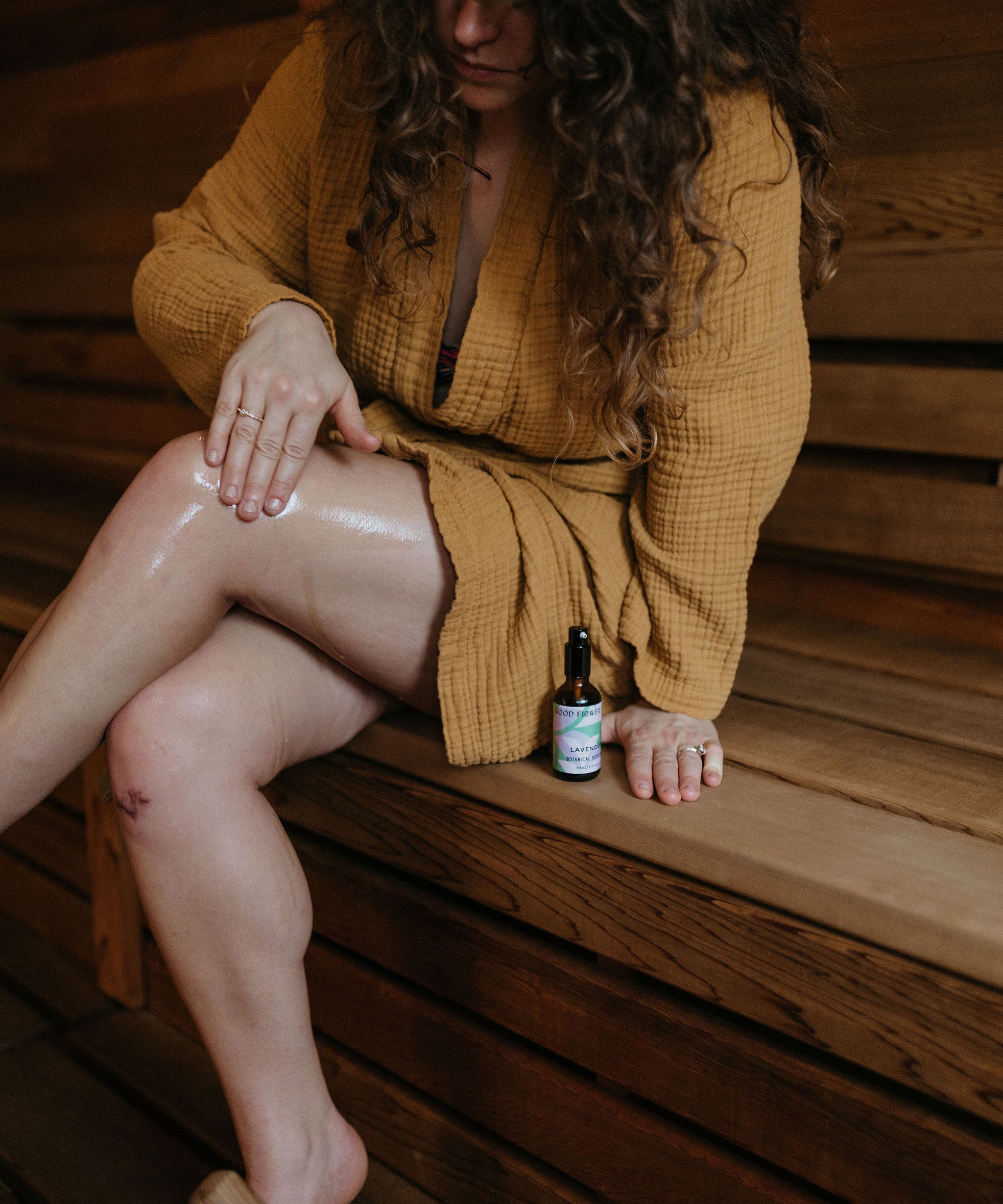 A woman in a mustard robe sits on a wooden bench, applying oil to her leg. A small bottle of oil is placed beside her on the bench.