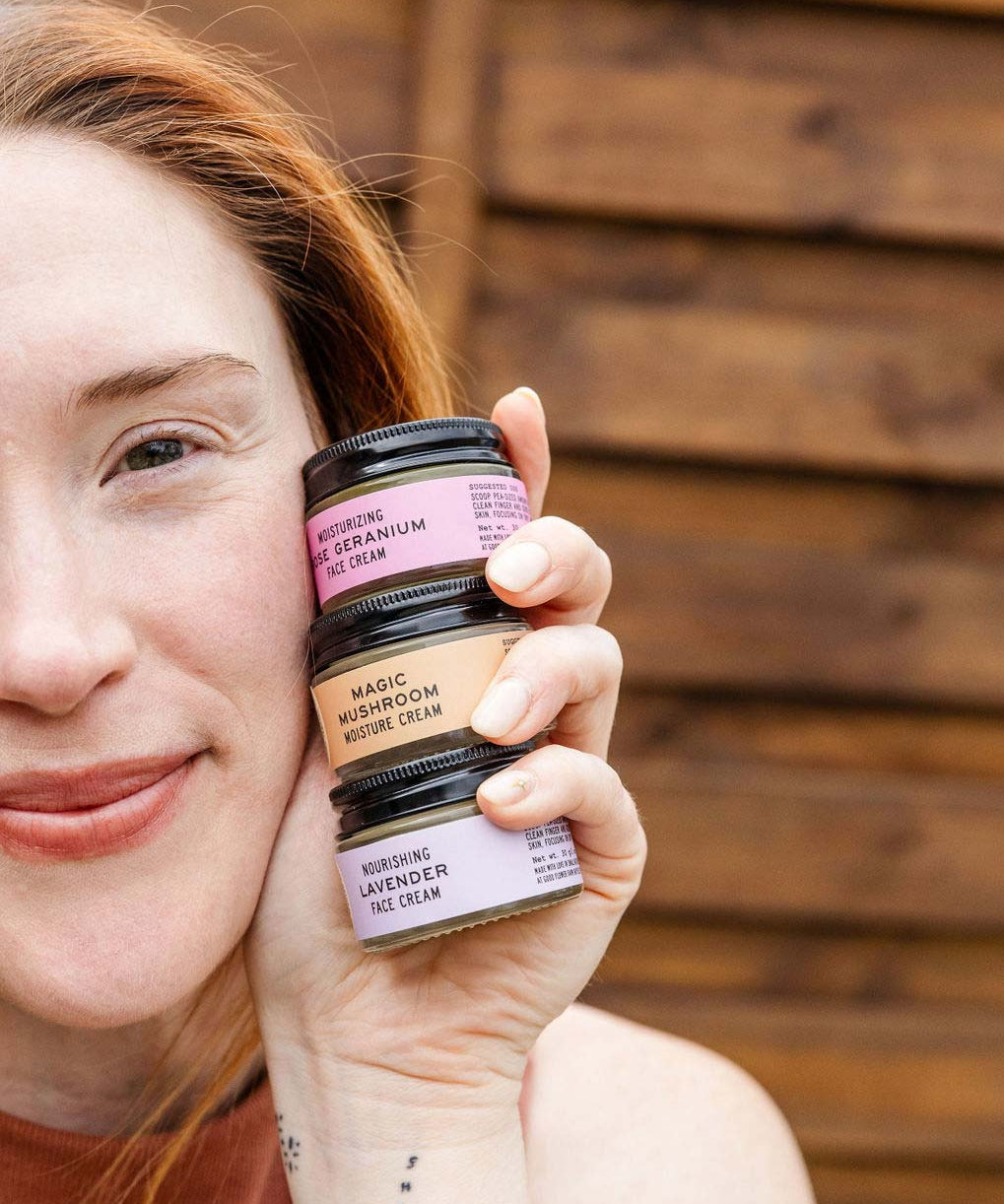 A woman with fair skin and auburn hair smiles, holding three small jars of face cream stacked by her cheek. The jars are labeled rose geranium, magic mushroom, and lavender. Wooden wall in background.