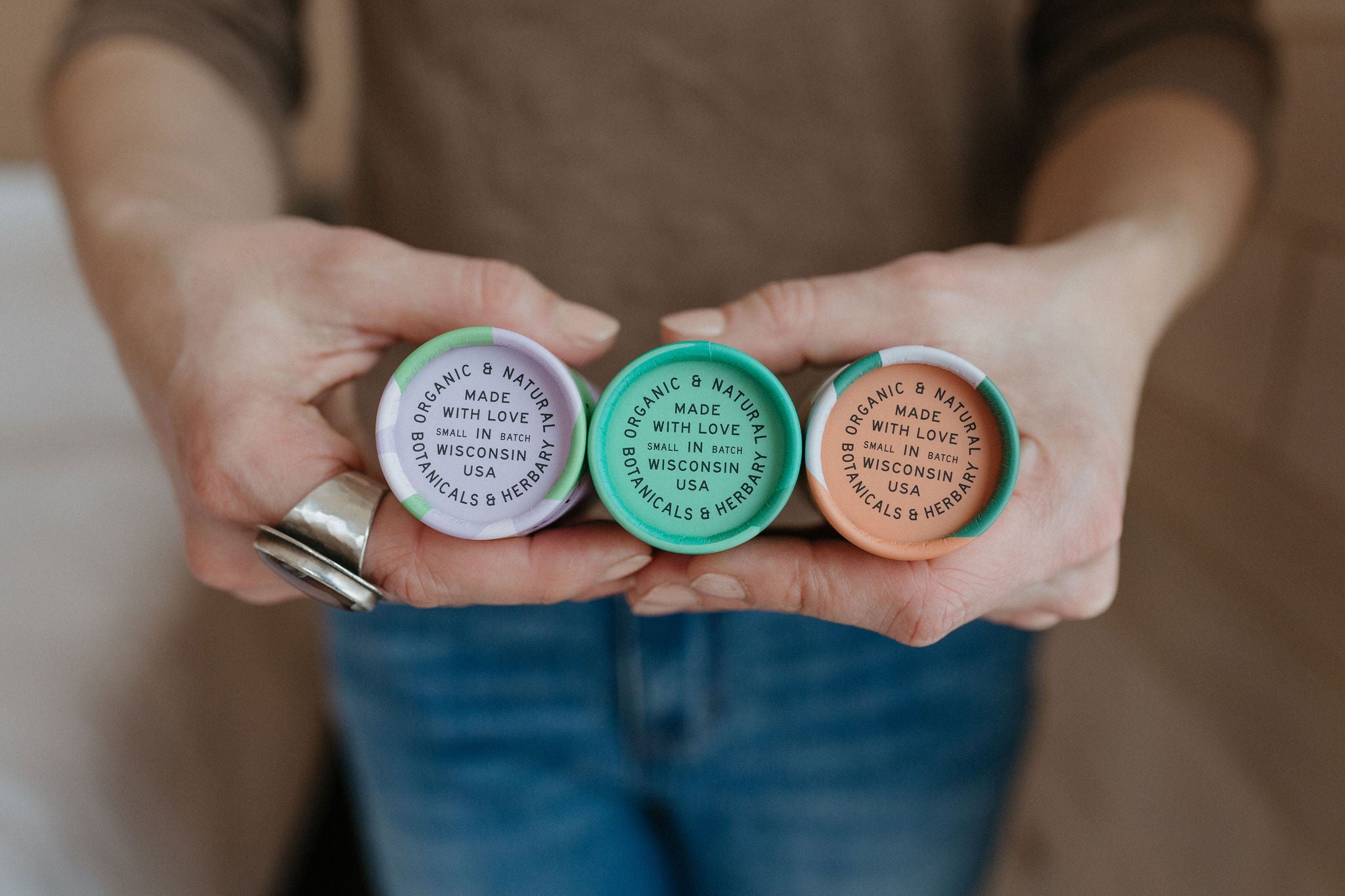 A person holding three colorful tubes with labels reading “Organic & Natural. Made with love in Wisconsin USA. Botanicals & Herbs.” The tubes are green, purple, and orange.