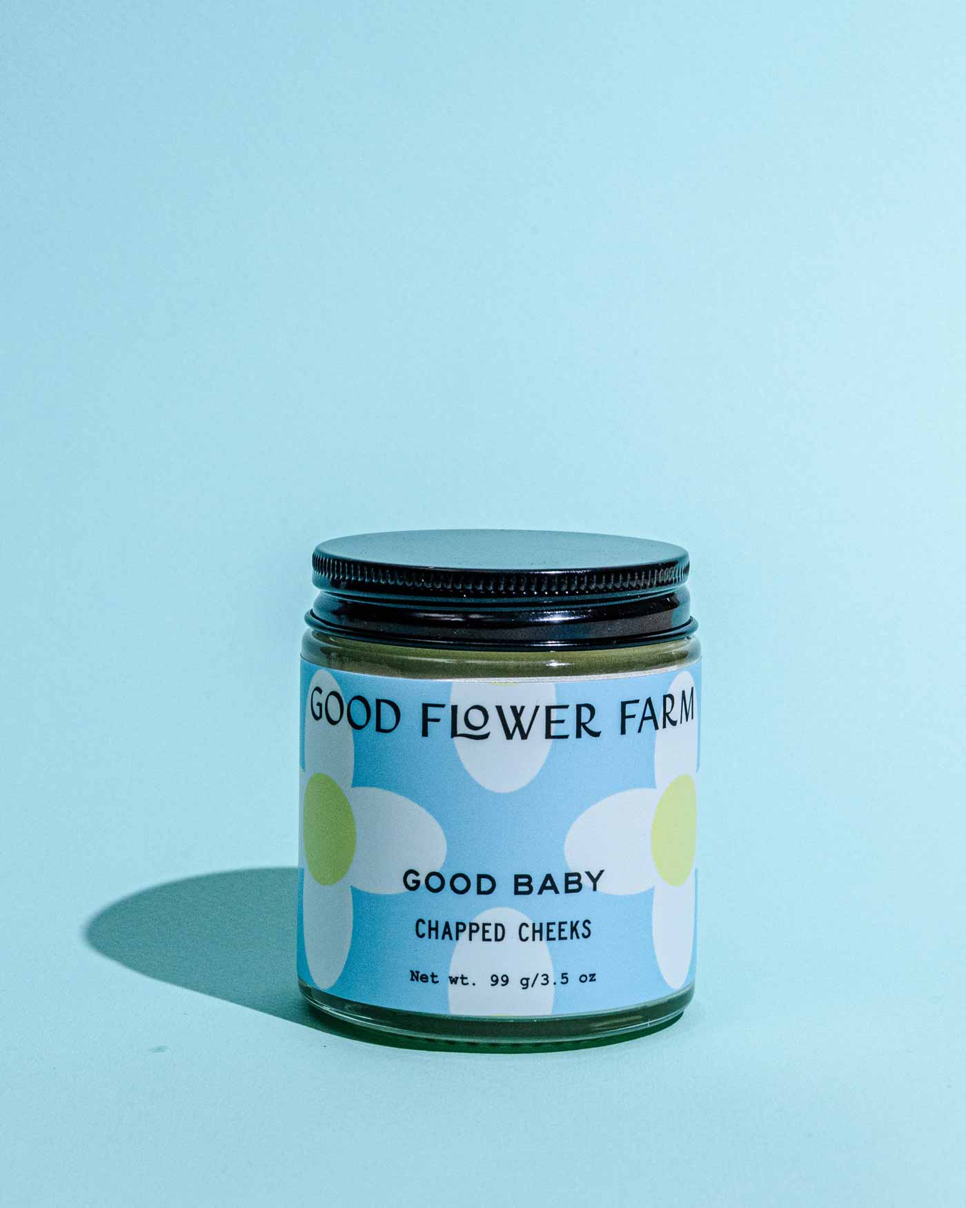 A glass jar labeled Good Flower Farm Good Baby Chapped Cheeks Diaper Balm with a floral design and black lid sits on a blue surface. The 99g (3.5oz) balm soothes delicate baby skin against a light blue background.