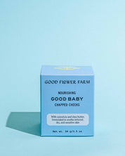 A blue box of Good Flower Farm’s Good Baby Chapped Cheeks Diaper Balm rests on a light blue background. The packaging highlights calendula and shea butter for nourishing, soothing diaper rash and sensitive baby skin.