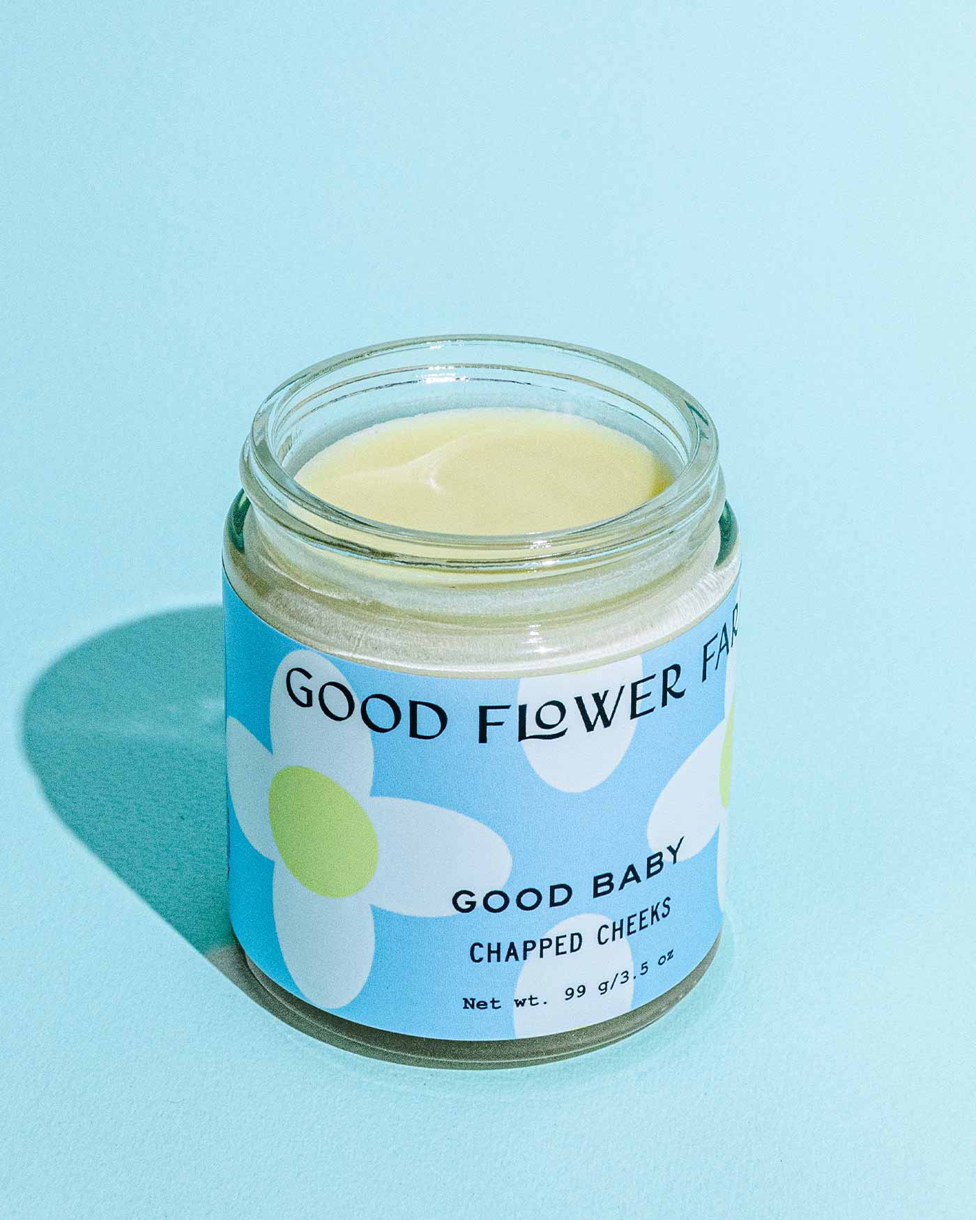 An open glass jar of Good Flower Farms TESTER — Good Baby Chapped Cheeks Diaper Balm sits on a light blue surface, showcasing a simple flower design and highlighting soothing calendula on the label.