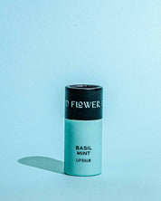 A cylindrical tube of Good Flower Farms TESTER — Basil Mint Lip Organic Lip Balm stands upright against a light blue background, with a black cap and light green body. The label reads Basil Mint Lip Balm. A soft shadow falls to the left.