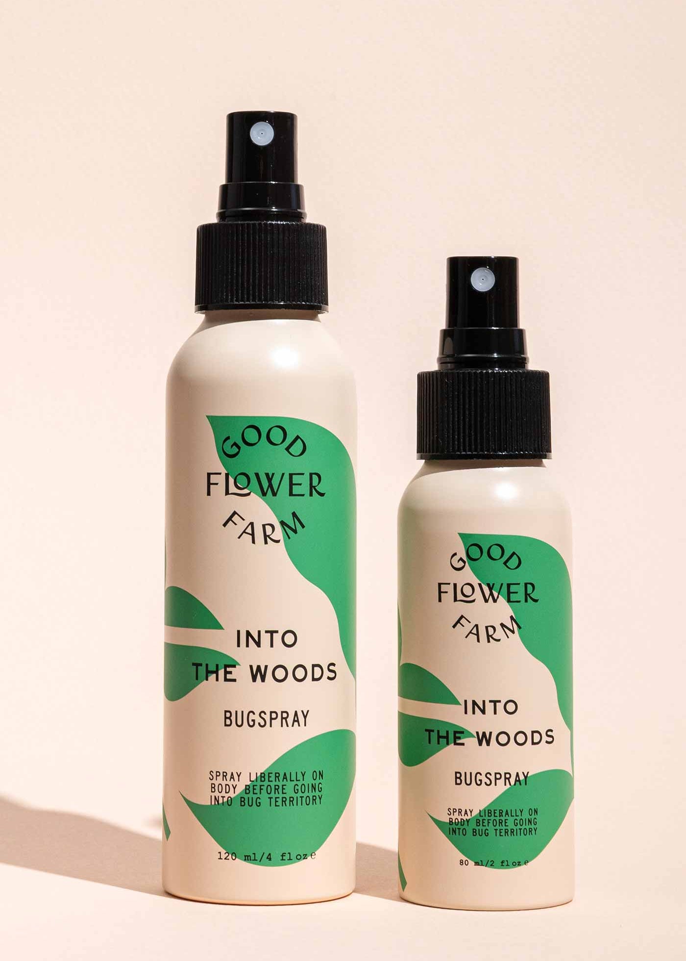 Two Good Flower Farm Into the Woods Natural Bugsprays, one large and one small, with green and cream labels, stand side by side on a beige background—the smaller is crafted with essential oils.
