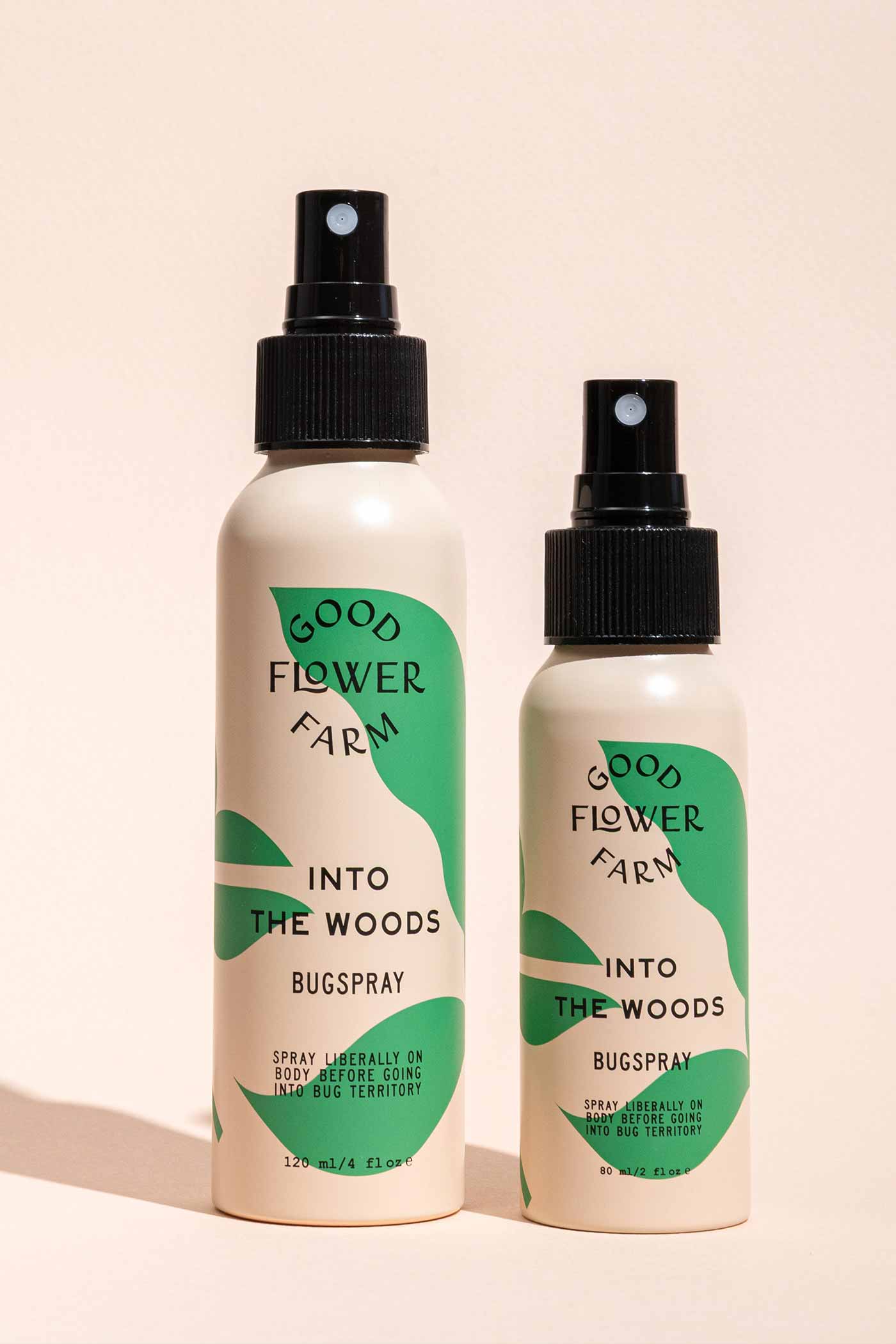 Two Good Flower Farm Into the Woods Natural Bugsprays, one large and one small, with green and cream labels, stand side by side on a beige background—the smaller is crafted with essential oils.