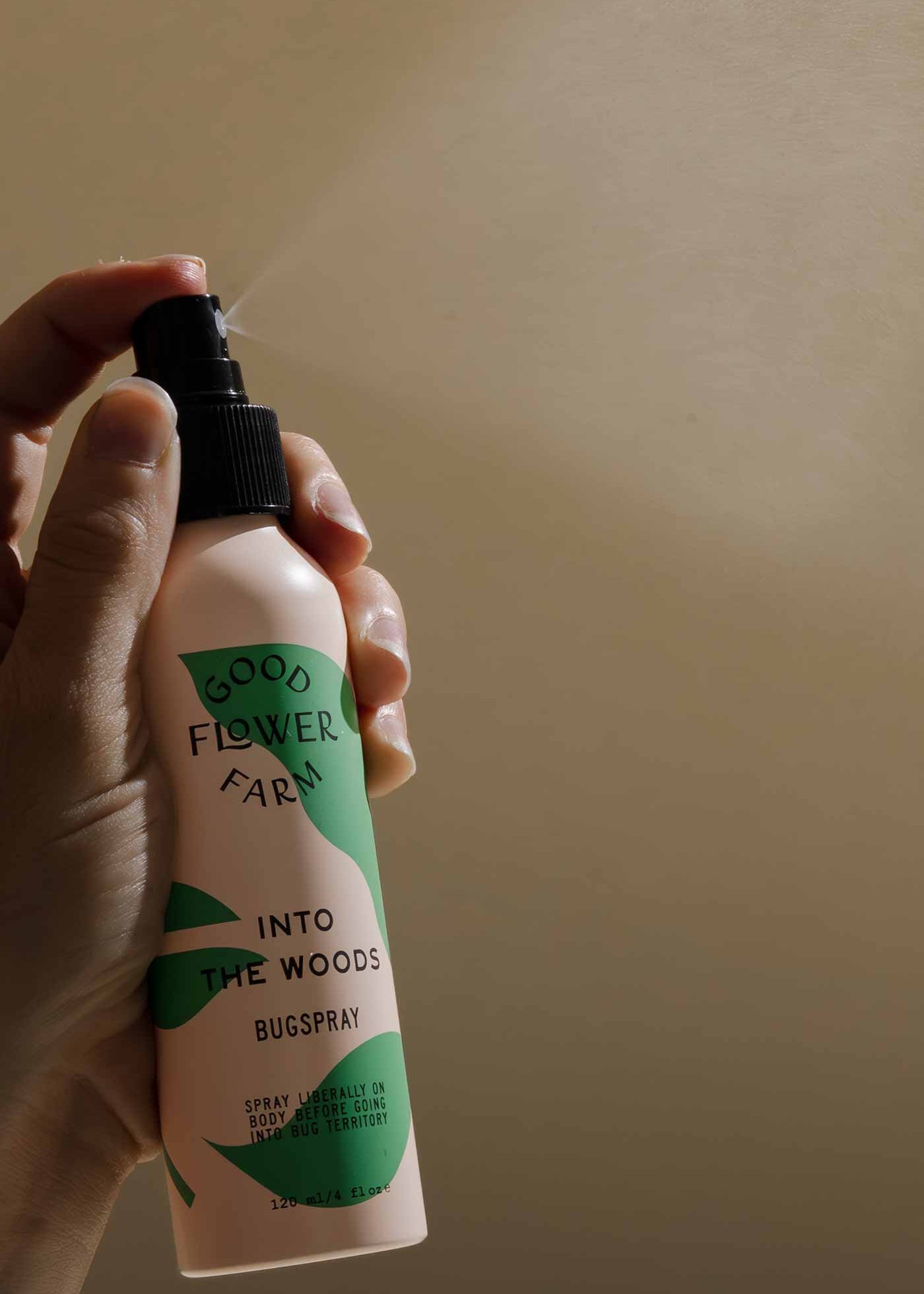 A hand holds a bottle of Good Flower Farm Into the Woods Natural Bugspray, an organic insect repellent with essential oils, set against a softly lit beige background.