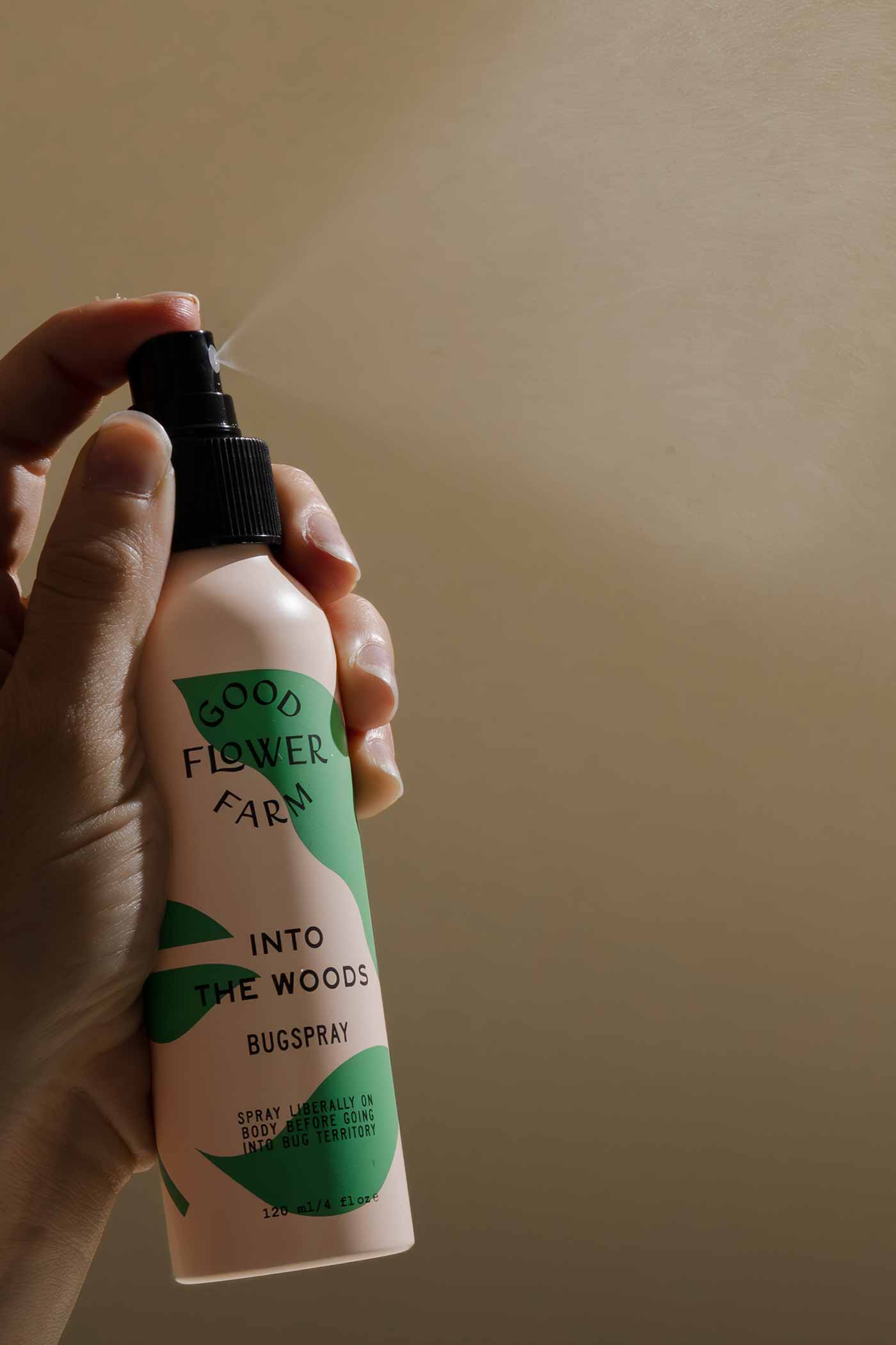 A hand holds a bottle of Good Flower Farm Into the Woods Natural Bugspray, an organic insect repellent with essential oils, set against a softly lit beige background.
