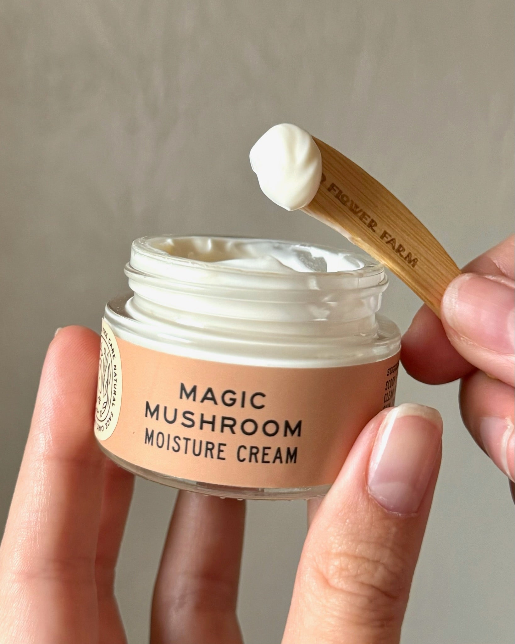 A hand holds a jar of Good Flower Farms Magic Mushroom Moisture Cream, while another hand displays a dollop of this adaptogenic mushroom skincare cream on a spatula above the open jar.