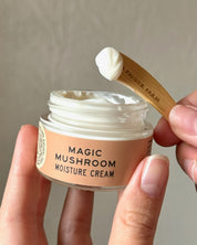 A hand holds a jar of Good Flower Farms Magic Mushroom Moisture Cream, while another hand displays a dollop of this adaptogenic mushroom skincare cream on a spatula above the open jar.