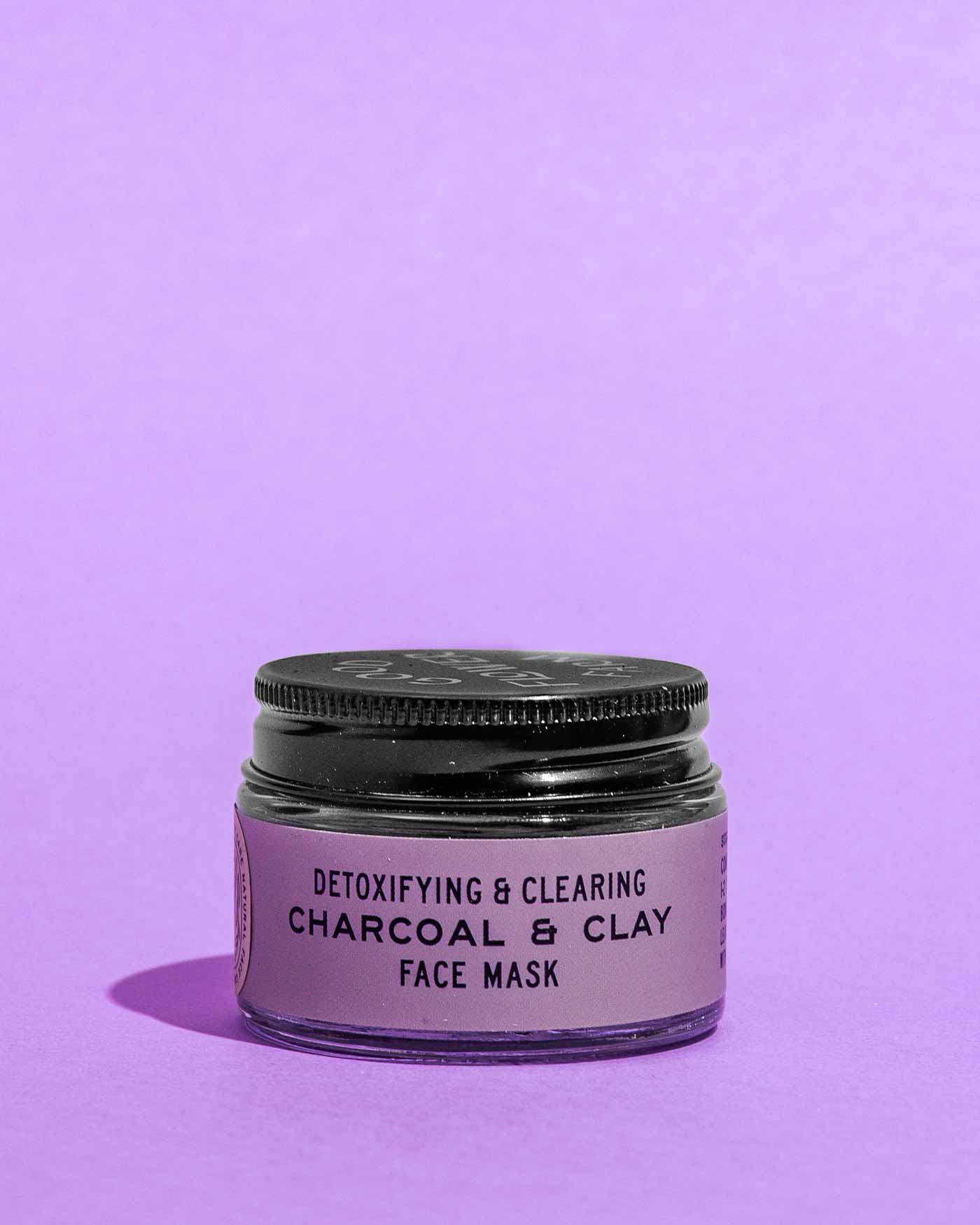 A small jar of Good Flower Farm Charcoal & Clay Botanical Face Mask with a black lid and purple label, featuring bentonite clay, sits on a purple surface against a matching purple background.
