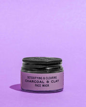 A small jar of Good Flower Farm Charcoal & Clay Botanical Face Mask with a black lid and purple label, featuring bentonite clay, sits on a purple surface against a matching purple background.