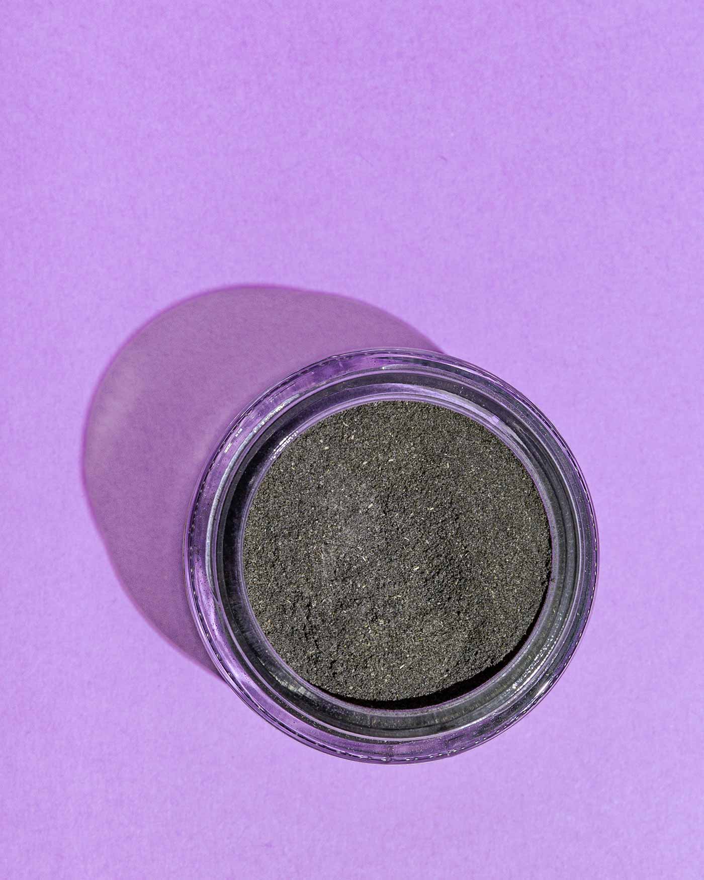 A jar of Good Flower Farm’s Charcoal & Clay Botanical Face Mask, filled with fine charcoal powder, rests on a purple surface with its shadow cast to the left.