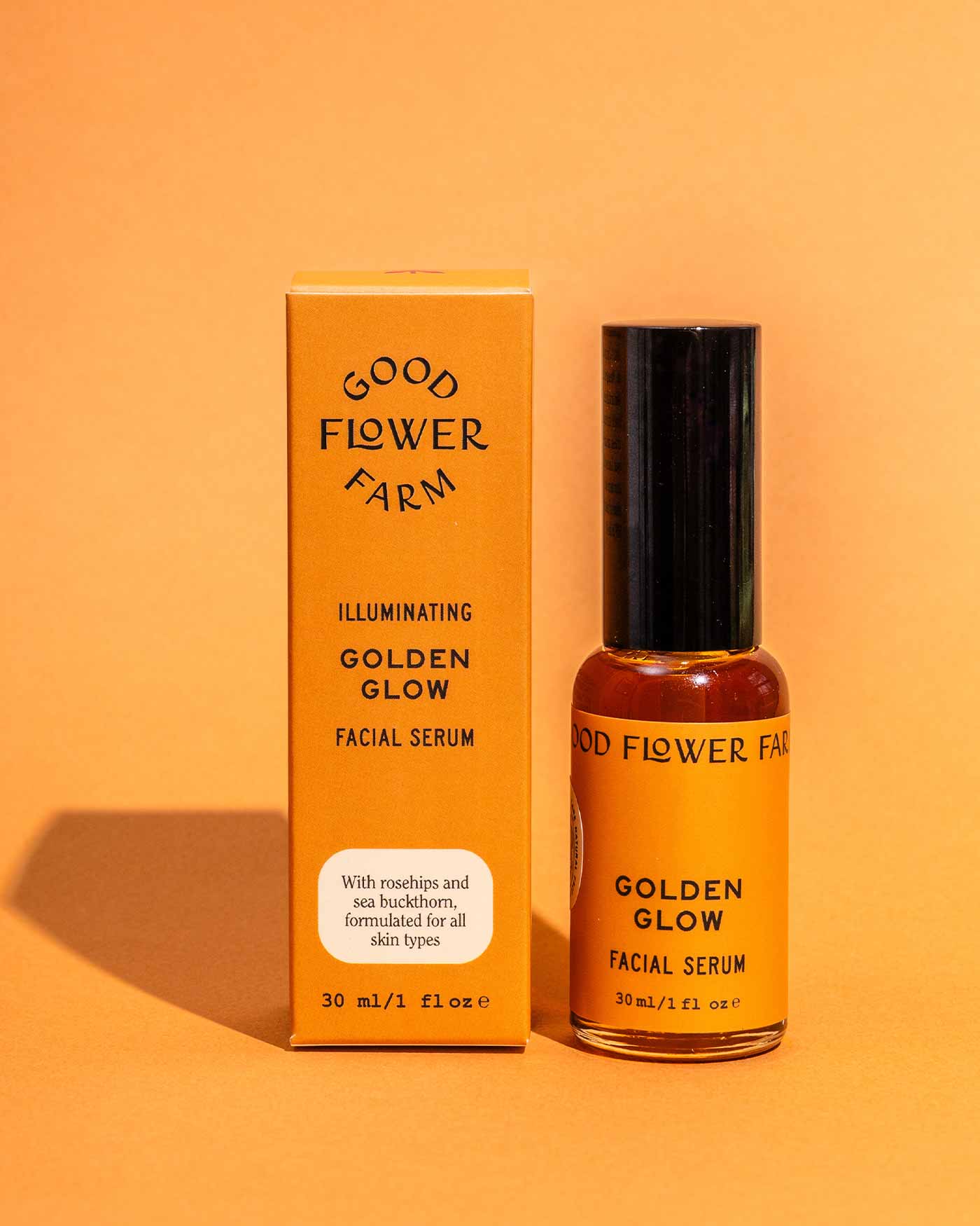 A bottle and box of Good Flower Farm Golden Glow Facial Serum are shown on an orange surface. The yellow packaging with black text highlights rosehips and sea buckthorn for a natural glow and hydrated skin.