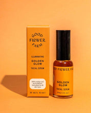 A bottle and box of Good Flower Farm Golden Glow Facial Serum are shown on an orange surface. The yellow packaging with black text highlights rosehips and sea buckthorn for a natural glow and hydrated skin.