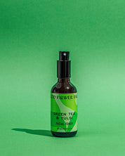 A brown bottle of Good Flower Farm Green Tea & Tulsi Facial Toner with a black spray cap sits against a green background. The green label displays the product name and highlights soothing ingredients: green tea and tulsi.