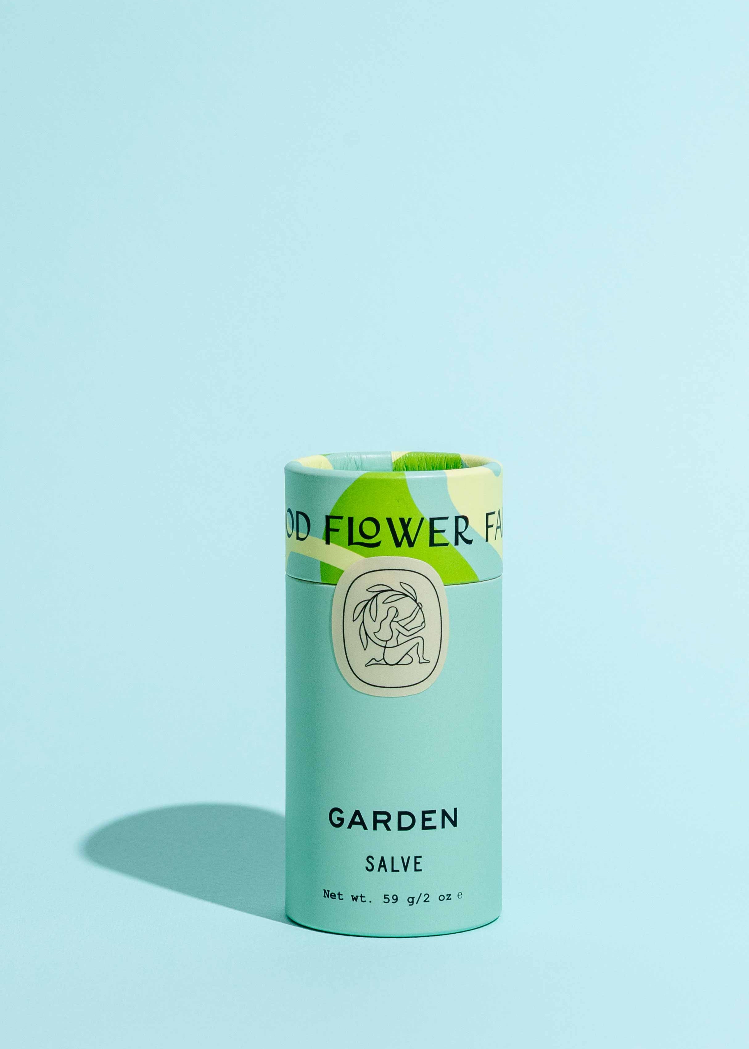 A cylindrical Garden Salve by Good Flower Farm stands upright on a light blue surface with a matching background, featuring light blue packaging, green accents, and an illustrated logo.