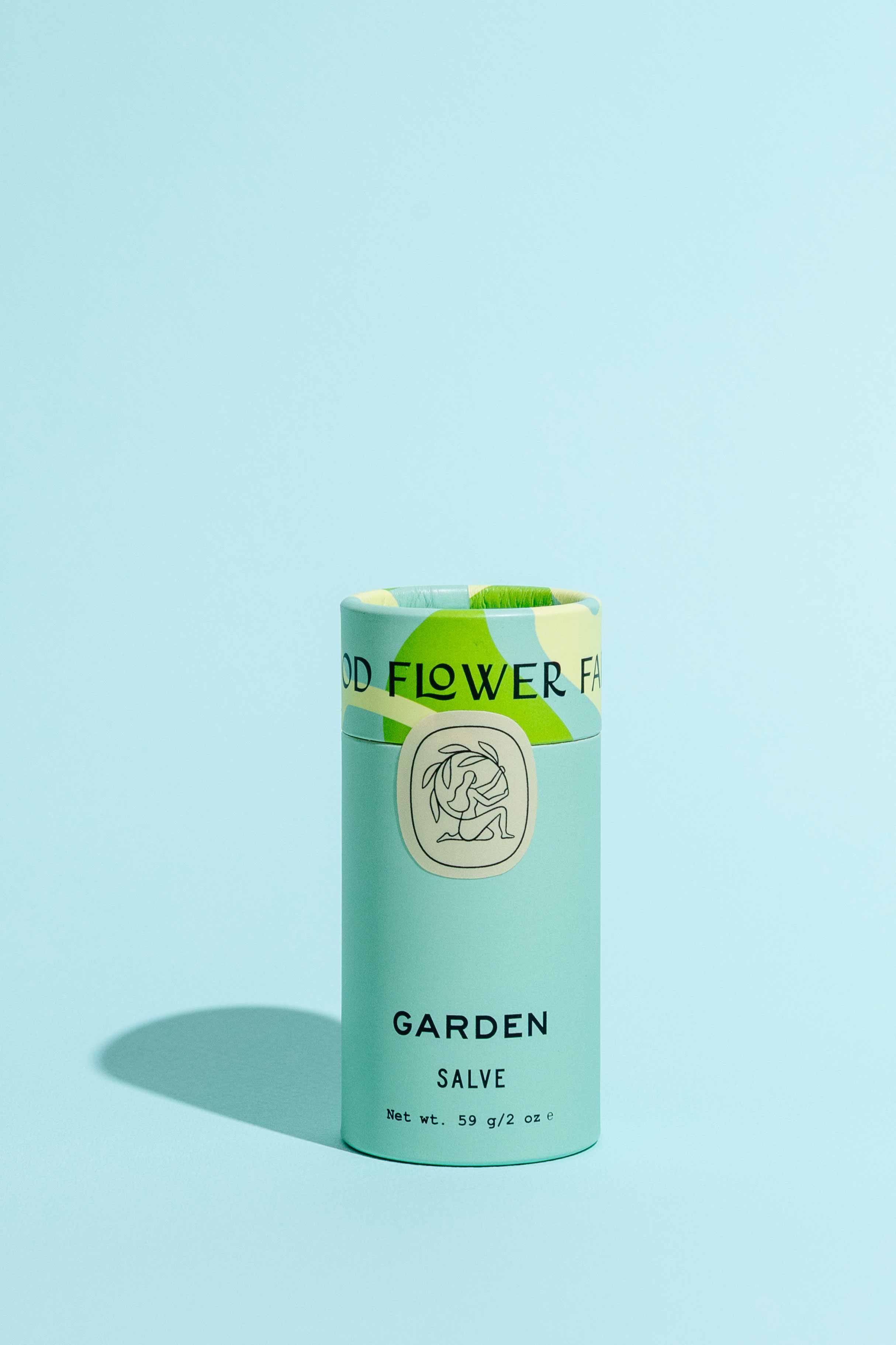 A cylindrical Garden Salve by Good Flower Farm stands upright on a light blue surface with a matching background, featuring light blue packaging, green accents, and an illustrated logo.
