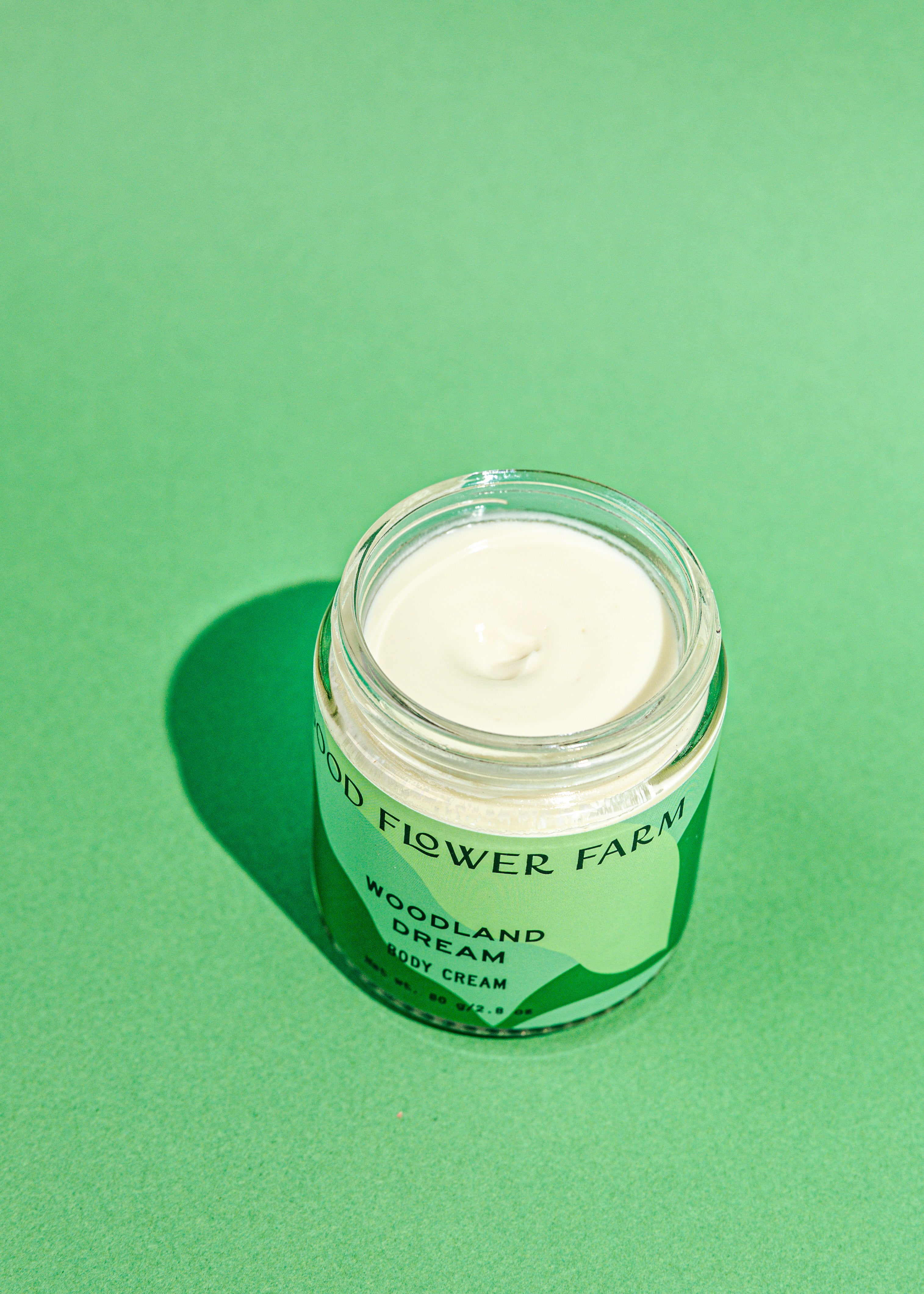 An open glass jar of TESTER — Woodland Dream Organic Body Cream by Good Flower Farm sits on a green background, revealing a swirl of shea butter and Douglas Fir hydrosol-infused cream inside.