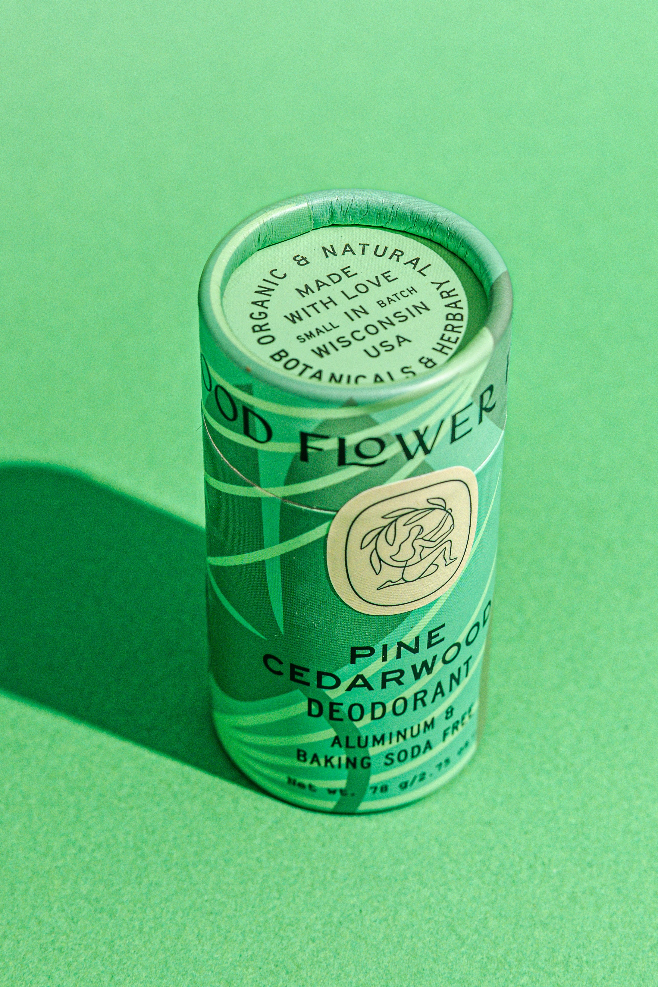 A cylindrical tube of Good Flower Farm Pine Cedarwood Deodorant sits on a green surface. The label notes it is organic, aluminum- and baking soda-free, and packaged in a biodegradable, plastic-free tube.