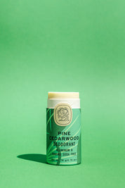 A Good Flower Farm Pine Cedarwood Deodorant Plastic-Free Tube stands upright on a solid green background, its green and cream biodegradable packaging emphasizing an aluminum- and baking soda-free formula.