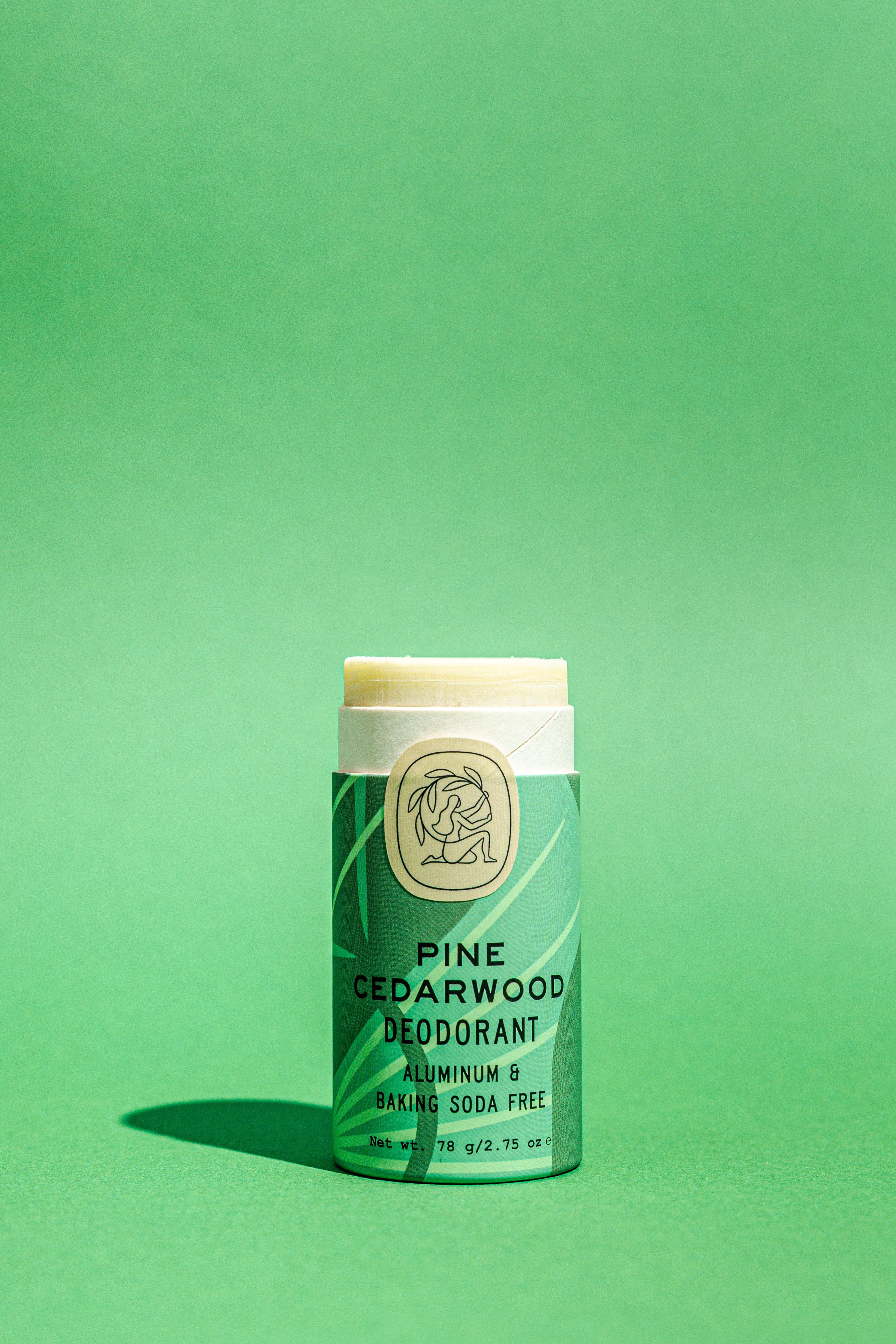 A Good Flower Farm Pine Cedarwood Deodorant Plastic-Free Tube stands upright on a solid green background, its green and cream biodegradable packaging emphasizing an aluminum- and baking soda-free formula.