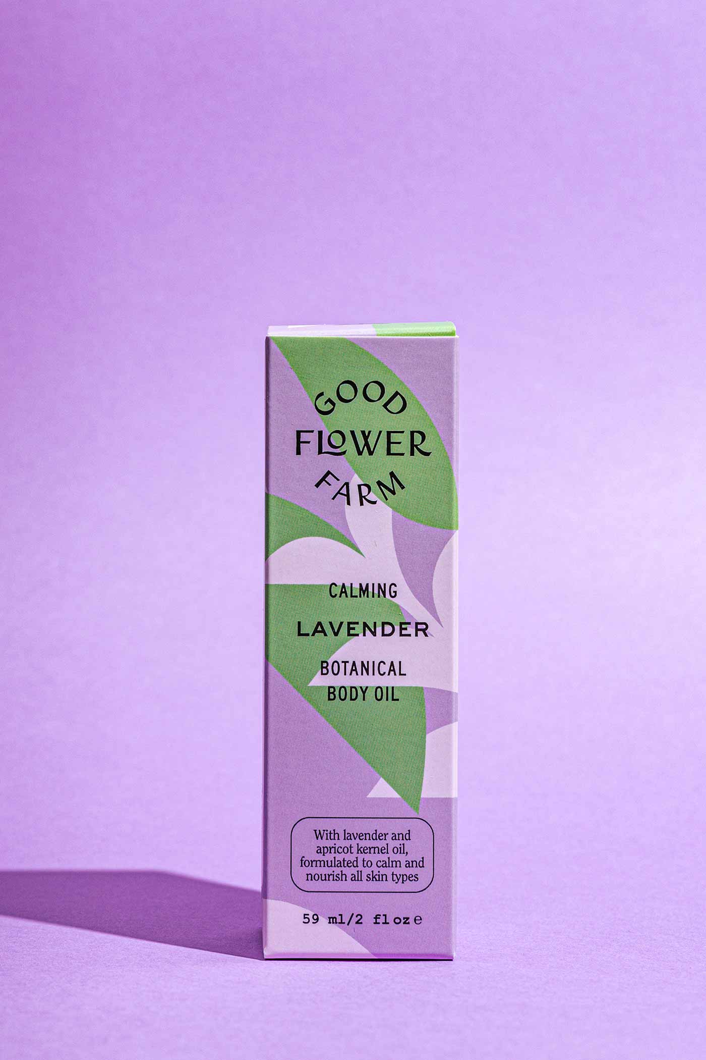 A rectangular box of Good Flower Farm Lavender Botanical Body Oil stands upright on a purple surface with a matching background. The elegant purple and green packaging highlights this soothing body oil with hemp seed oil.