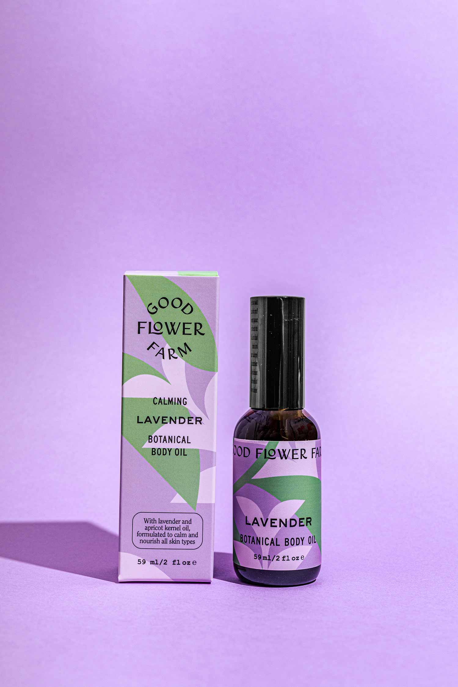 A bottle of Good Flower Farm Lavender Botanical Body Oil sits beside its matching lavender and green box on a purple surface with a left-cast shadow—ideal for relaxation as a soothing massage oil.