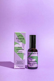 A bottle of Good Flower Farm Lavender Botanical Body Oil sits beside its matching lavender and green box on a purple surface with a left-cast shadow—ideal for relaxation as a soothing massage oil.