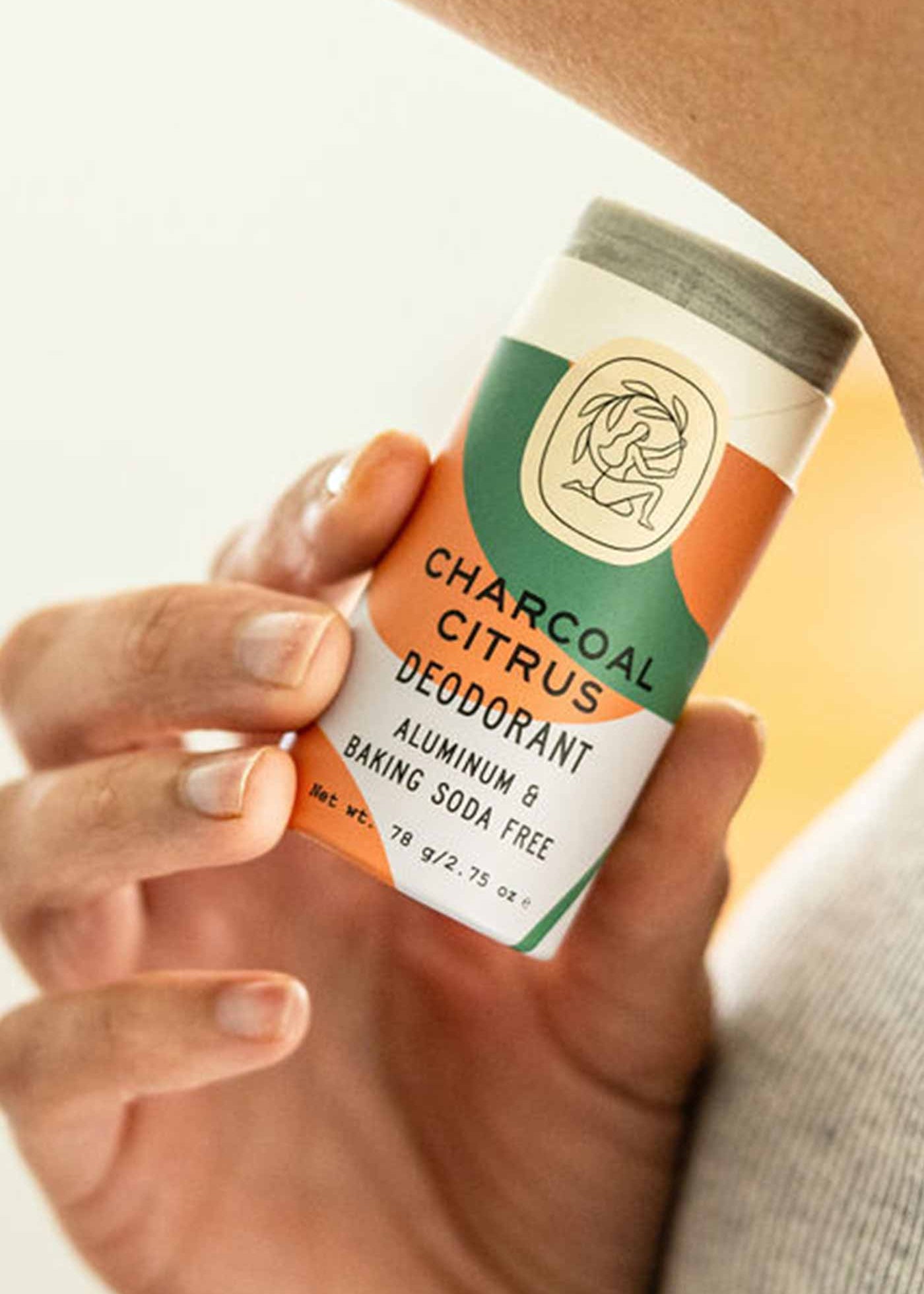 A hand holds a Good Flower Farm Charcoal Citrus Deodorant Plastic-Free Tube, made with essential oils and free from aluminum and baking soda, featuring a vibrant orange, green, and cream design.
