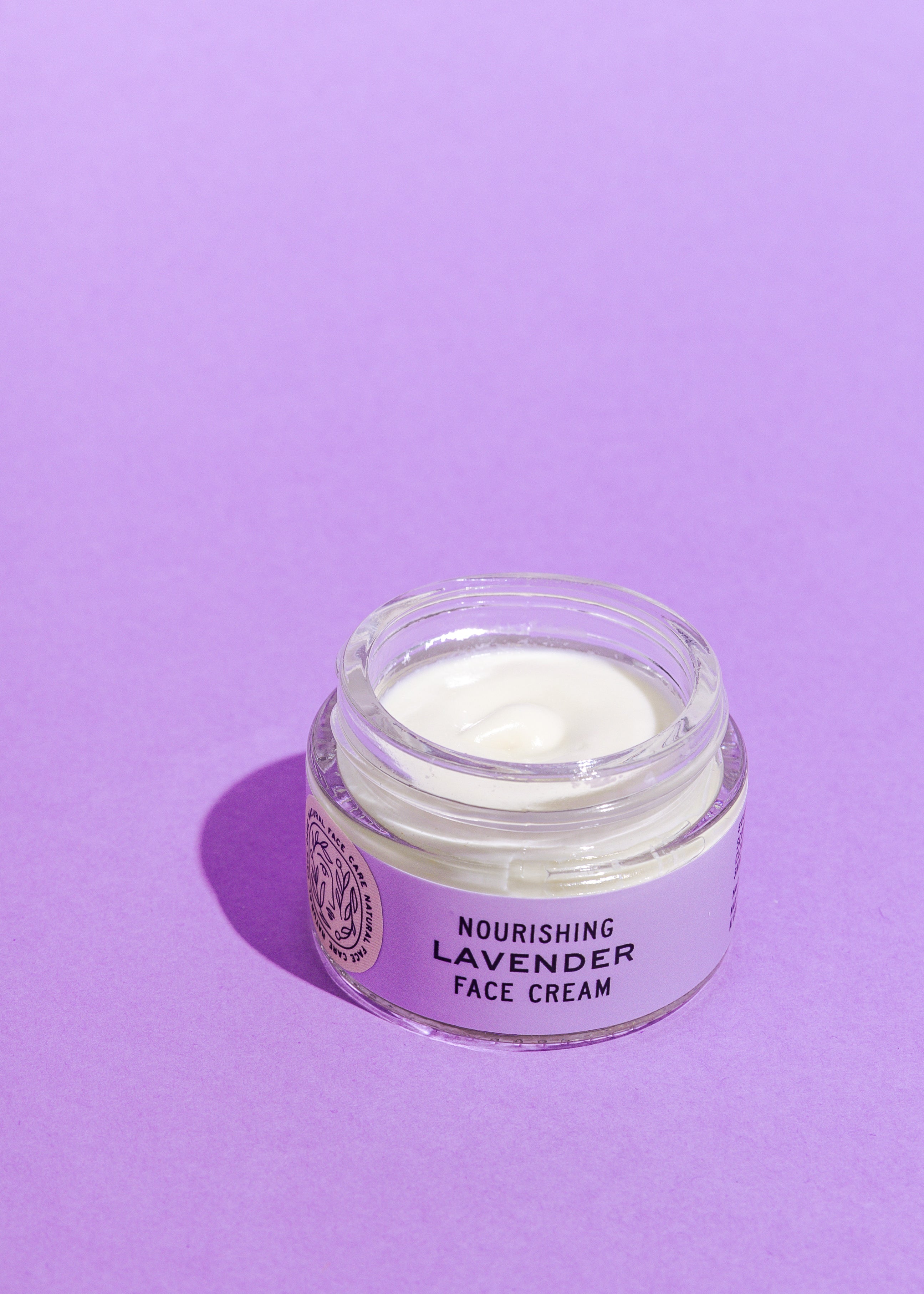 A glass jar of Good Flower Farm’s Lavender Face Cream sits open on a purple surface, revealing the creamy white, calendula-infused moisturizer inside.