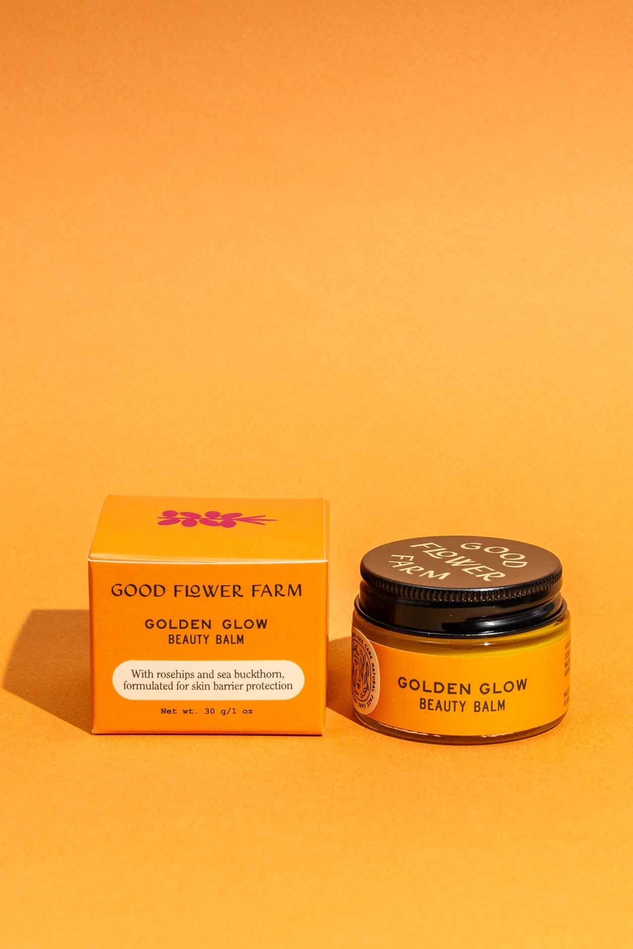 A jar and box of Good Flower Farm Golden Glow Beauty Balm, a skin repair balm, are shown on an orange background. The orange packaging features white text and purple flower graphics.