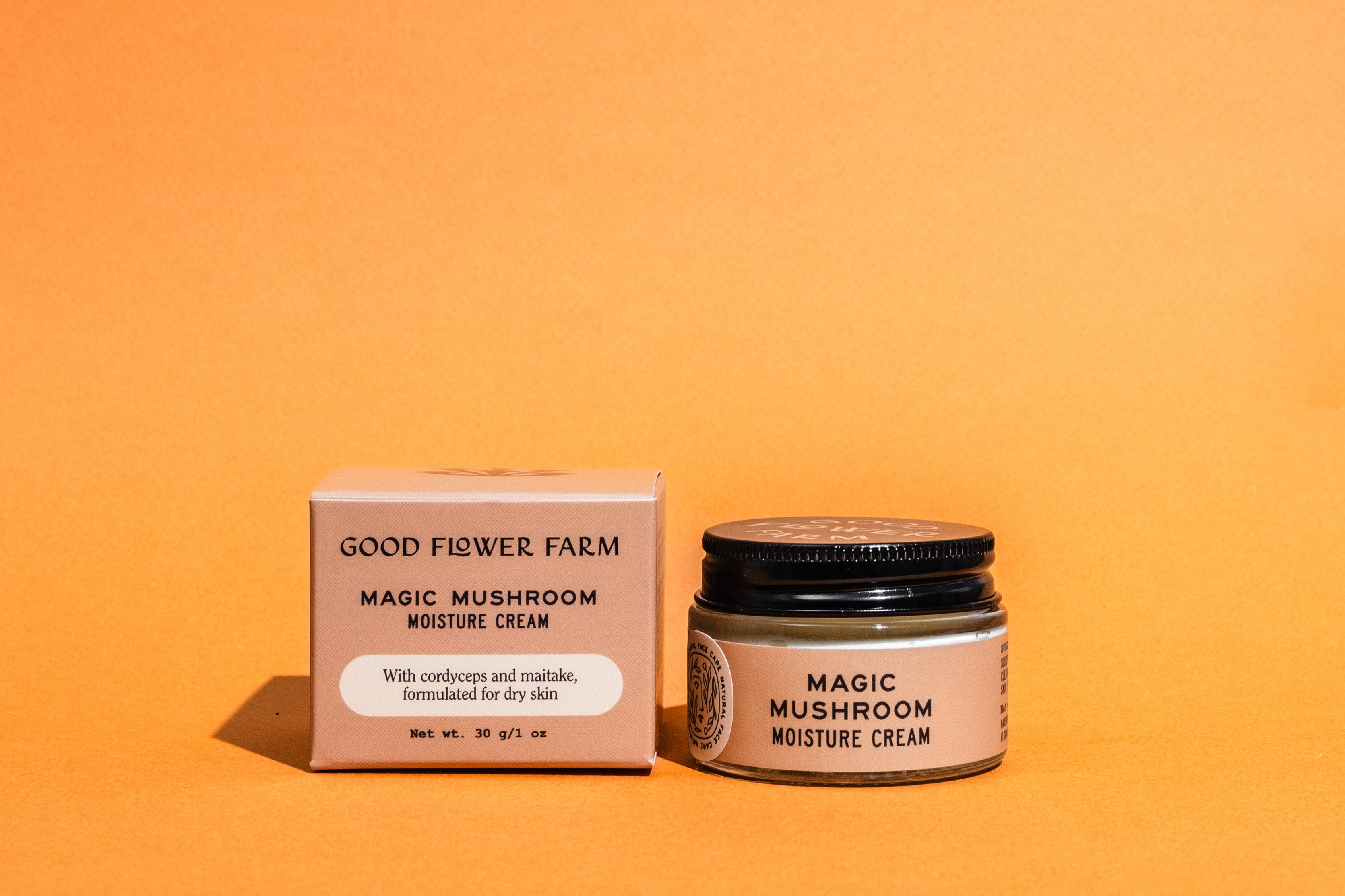 A jar and box of Good Flower Farm Magic Mushroom Moisture Cream sit on an orange background. This skincare is made for dry skin, featuring cordyceps oil and adaptogenic maitake, as highlighted on the packaging.
