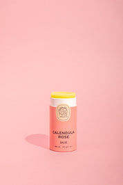 A tube of Good Flower Farm Calendula Rose Salve with a pink and cream label stands upright on a light pink background, the balm slightly exposed to showcase its soothing formula and eco-friendly packaging.