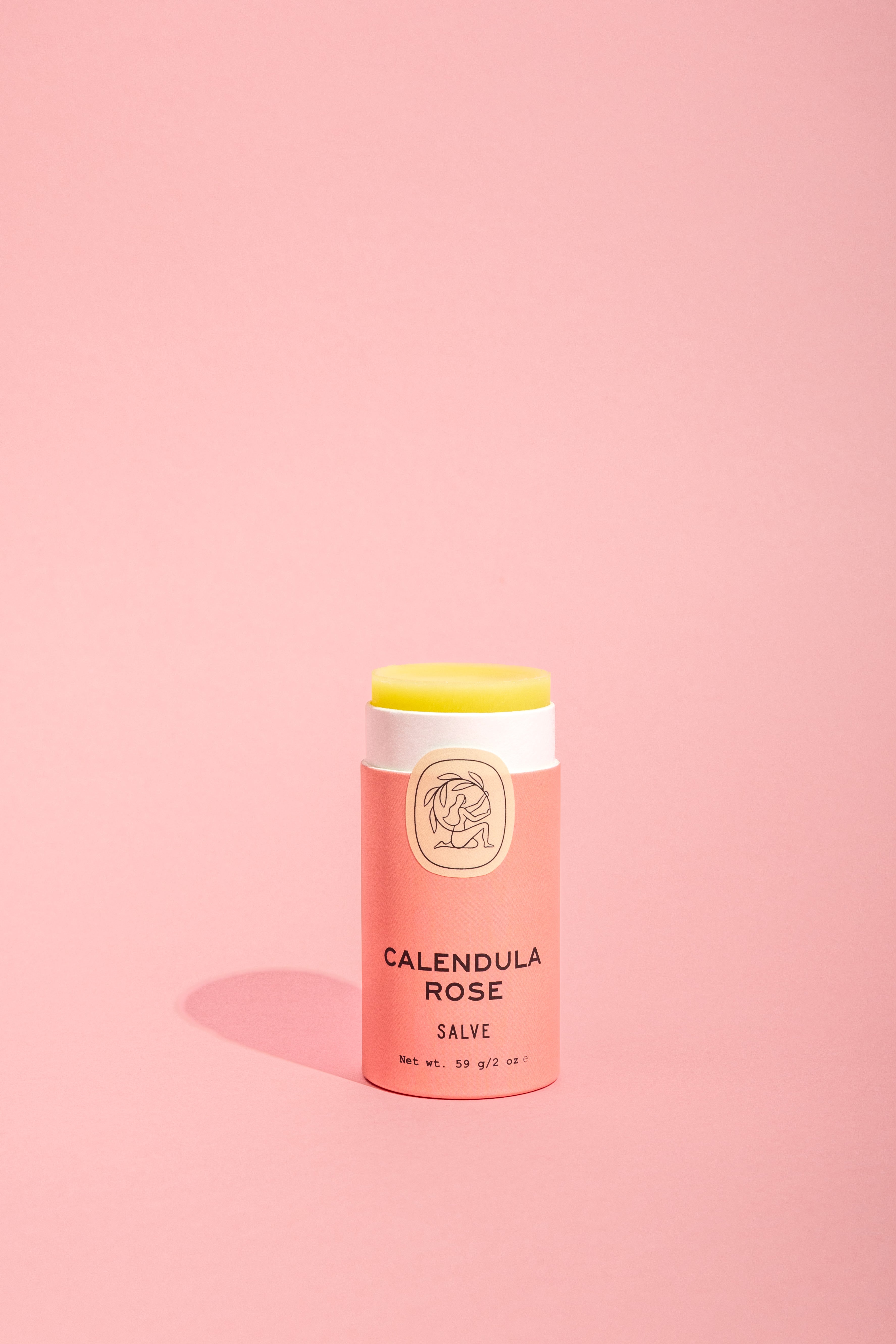 A tube of Good Flower Farm Calendula Rose Salve with a pink and cream label stands upright on a light pink background, the balm slightly exposed to showcase its soothing formula and eco-friendly packaging.