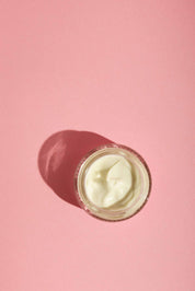 A small glass jar of Good Flower Farms Rose Geranium Face Cream sits on a pink background, white cream visible inside and casting a soft shadow to the left.