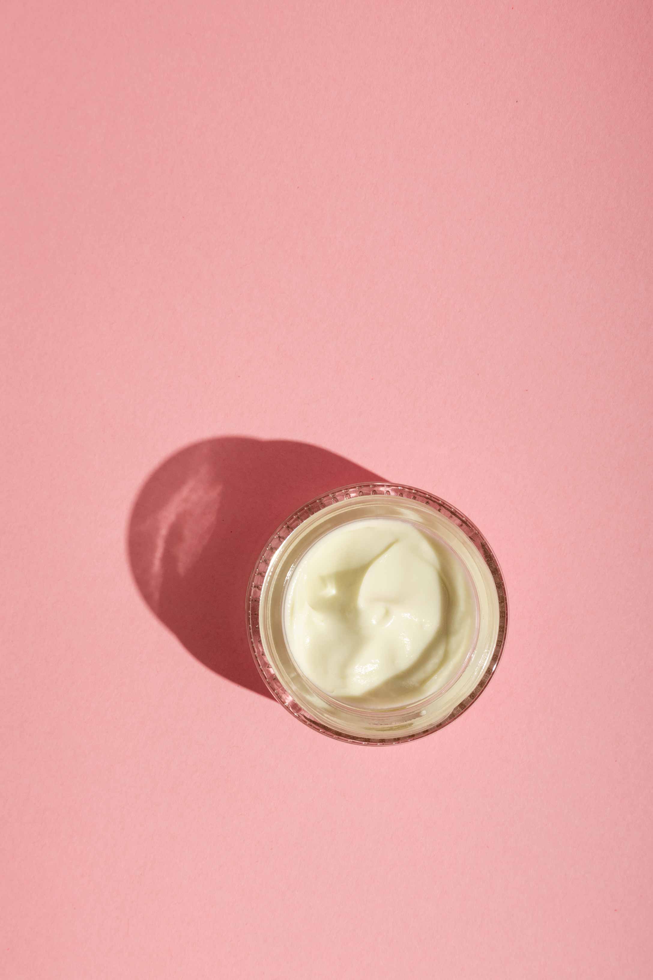 A small glass jar of Good Flower Farms Rose Geranium Face Cream sits on a pink background, white cream visible inside and casting a soft shadow to the left.