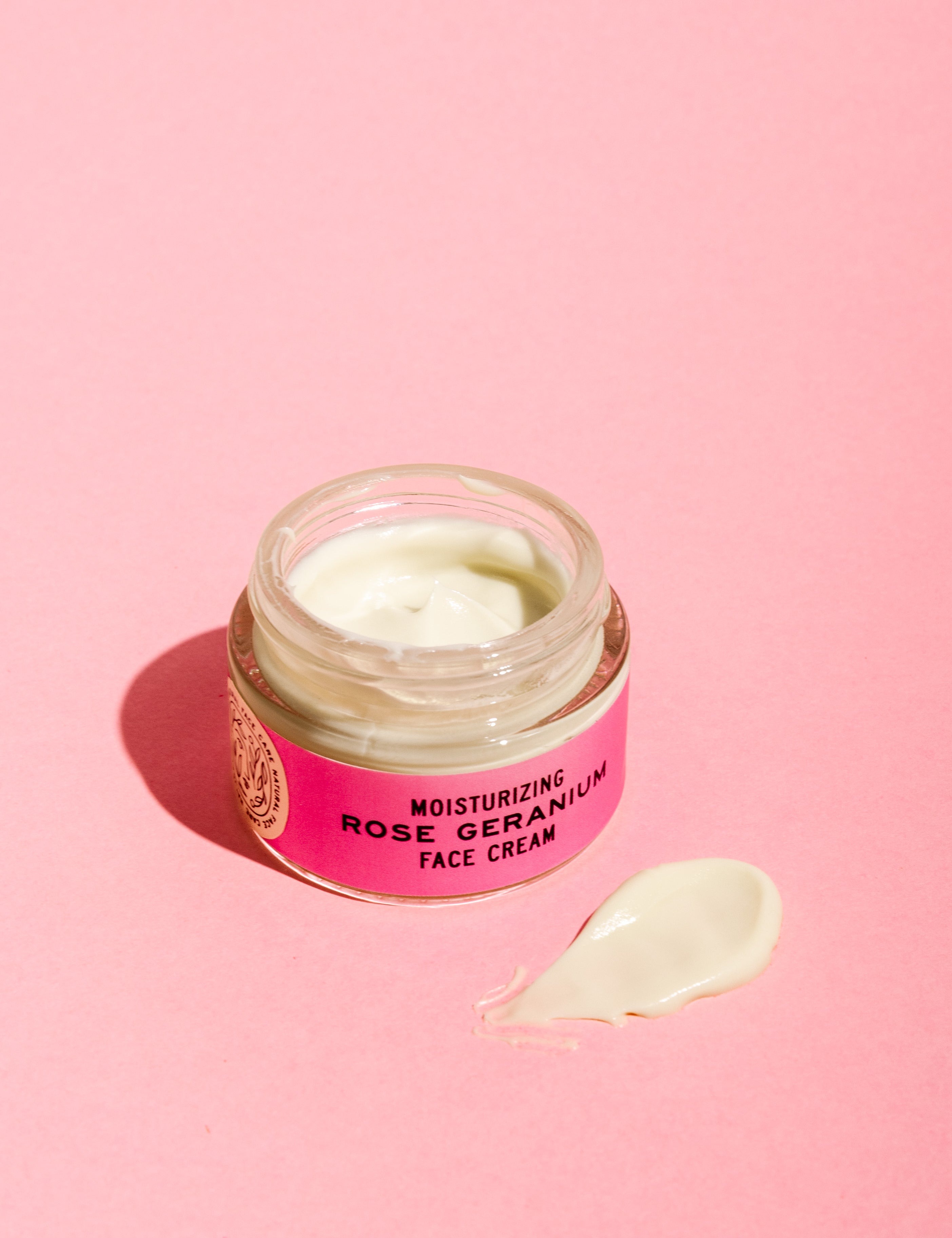 A jar of Good Flower Farm Rose Geranium Face Cream with soothing evening primrose oil sits open on a pink surface, its light-colored cream and dollop visible. The jar features a pink label.