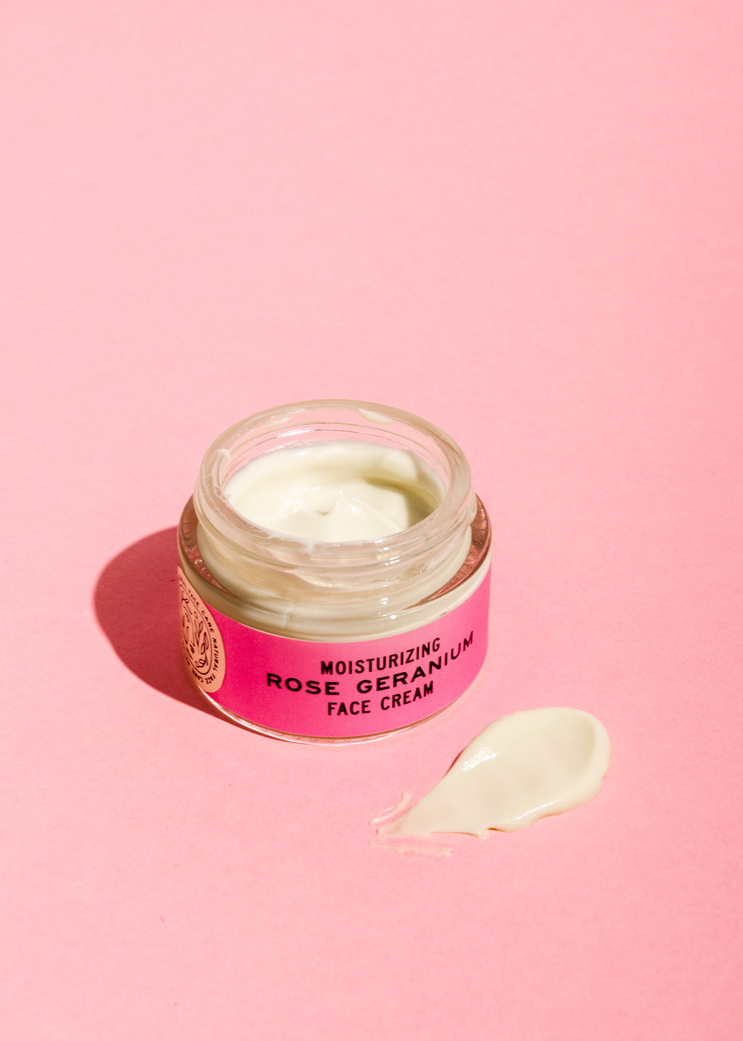 A jar of Good Flower Farm Rose Geranium Face Cream with soothing evening primrose oil sits open on a pink surface, its light-colored cream and dollop visible. The jar features a pink label.