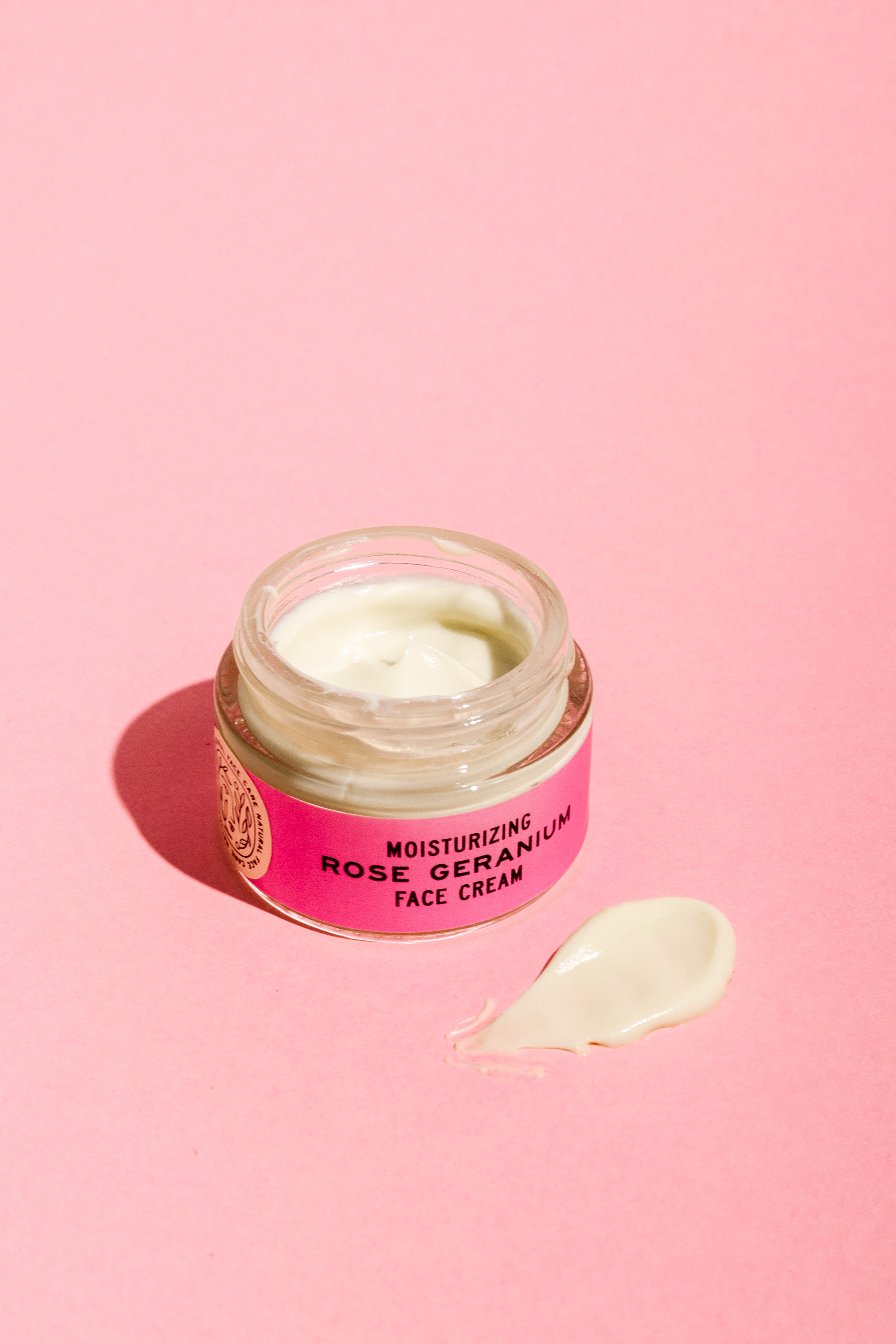 A jar of Good Flower Farm Rose Geranium Face Cream with soothing evening primrose oil sits open on a pink surface, its light-colored cream and dollop visible. The jar features a pink label.