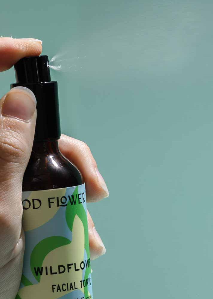 A hand sprays Good Flower Farm’s Wildflower Facial Tonic, releasing a fine mist of balancing toner with wild herbal flowers against a plain, light blue background.