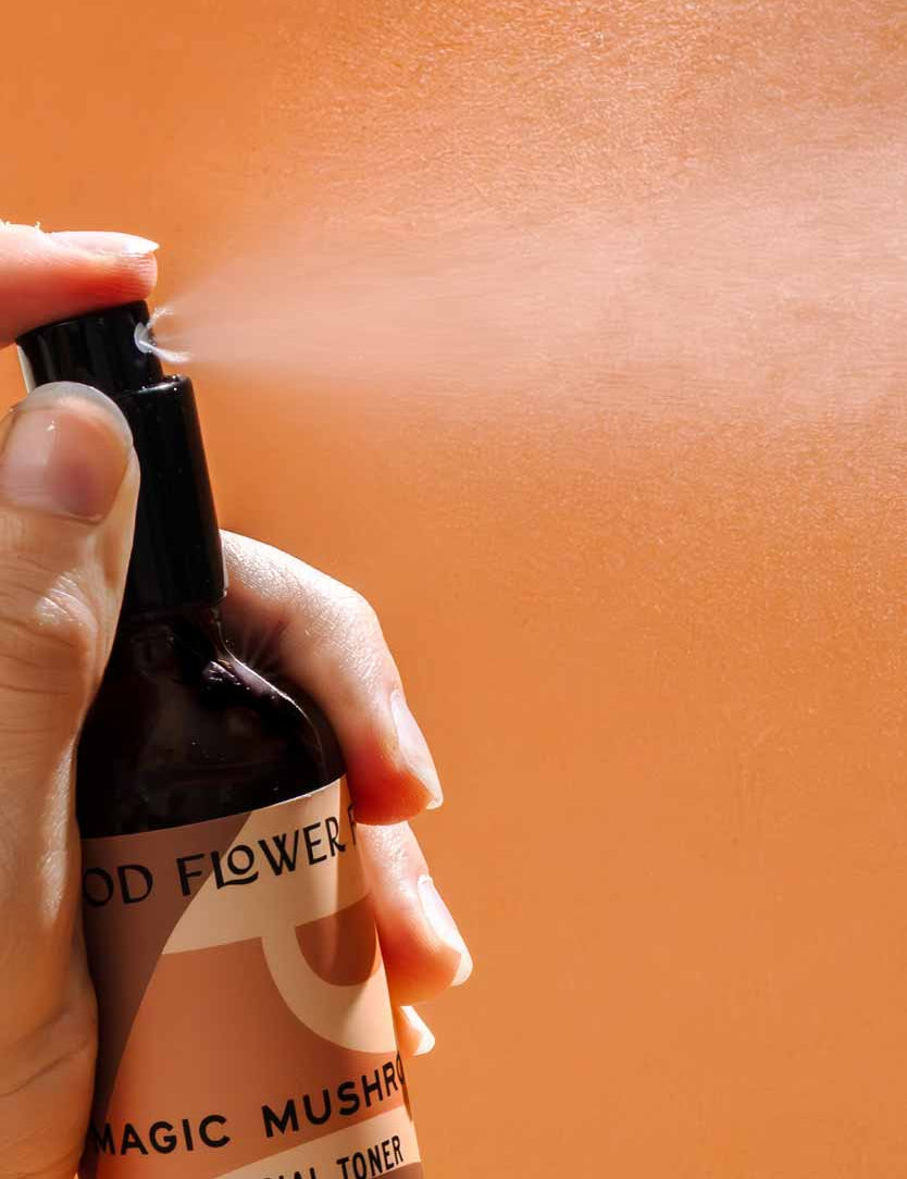 A hand sprays Good Flower Farm’s Magic Mushroom Facial Toner from a brown bottle labeled with its name, releasing a hydrating mist against an orange background.