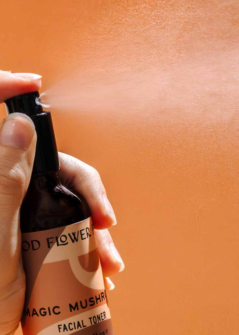 A hand sprays Good Flower Farm’s Magic Mushroom Facial Toner from a brown bottle labeled with its name, releasing a hydrating mist against an orange background.