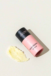 A pink tube of Good Flower Farm Raspberry Organic Lip Balm with a black cap rests on a light surface beside a small smear of the moisturizing, eco-friendly yellowish balm.