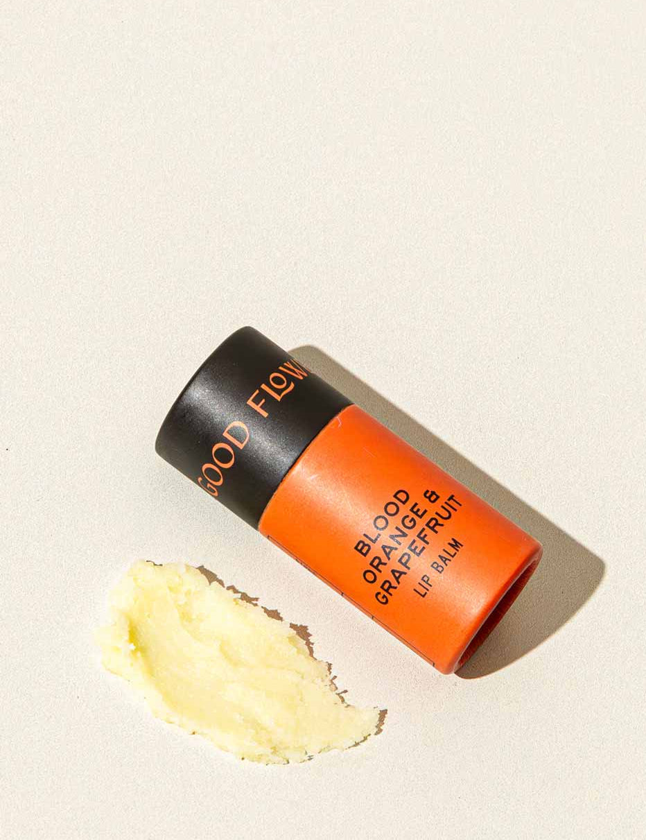 A tube of Good Flower Farm Blood Orange & Grapefruit Organic Lip Balm in orange and black rests on a light surface, beside a small smear of the moisturizing, eco-friendly balm.