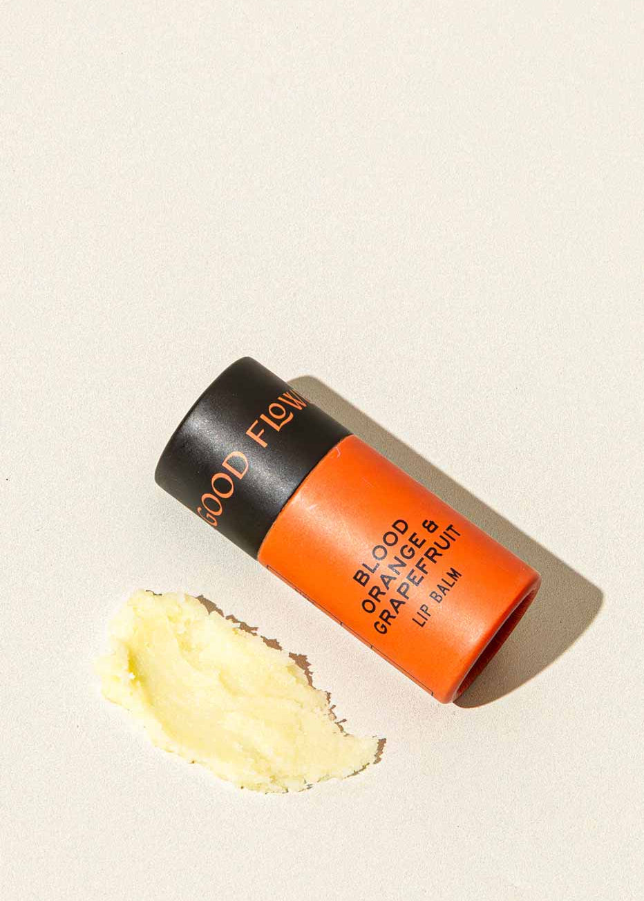 A tube of Good Flower Farm Blood Orange & Grapefruit Organic Lip Balm in orange and black rests on a light surface, beside a small smear of the moisturizing, eco-friendly balm.