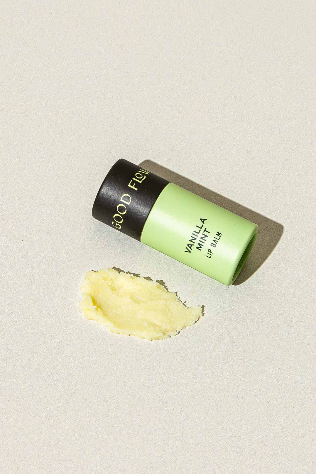 A green and black biodegradable tube of Good Flower Farms Vanilla Mint Organic Lip Balm lies on its side on a light surface, with a small smear of yellow coconut oil-infused balm spread nearby.
