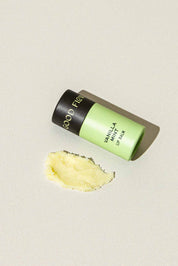 A green and black biodegradable tube of Good Flower Farms Vanilla Mint Organic Lip Balm lies on its side on a light surface, with a small smear of yellow coconut oil-infused balm spread nearby.