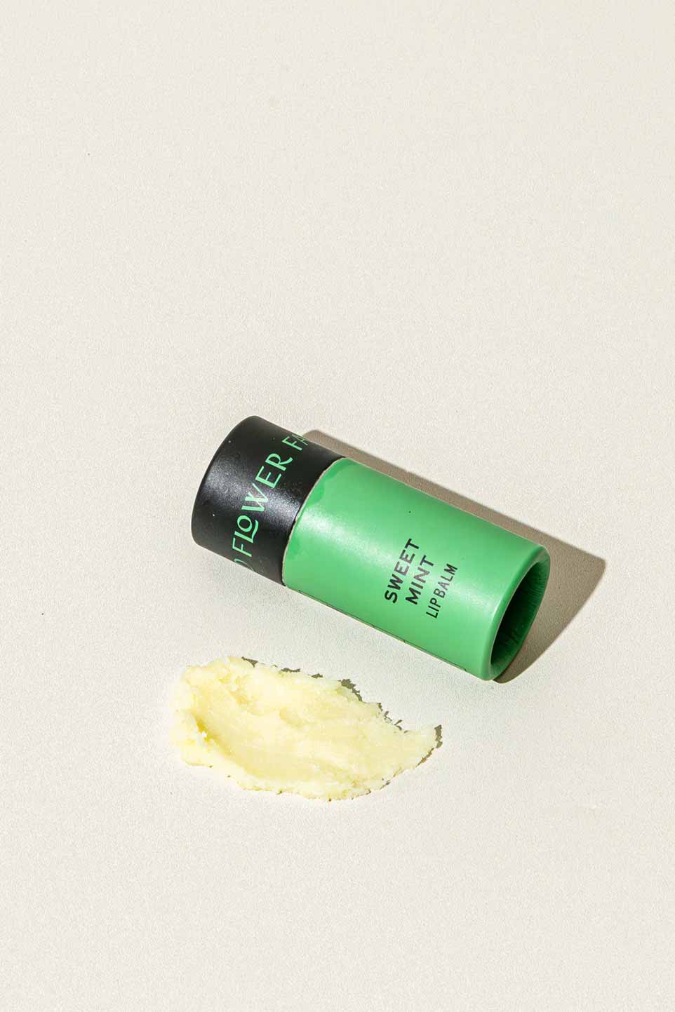 A green tube labeled “Sweet Mint Organic Lip Balm” by Good Flower Farm rests on its side on a light surface, with a small amount of yellowish balm spread in front of this eco-friendly lip balm.