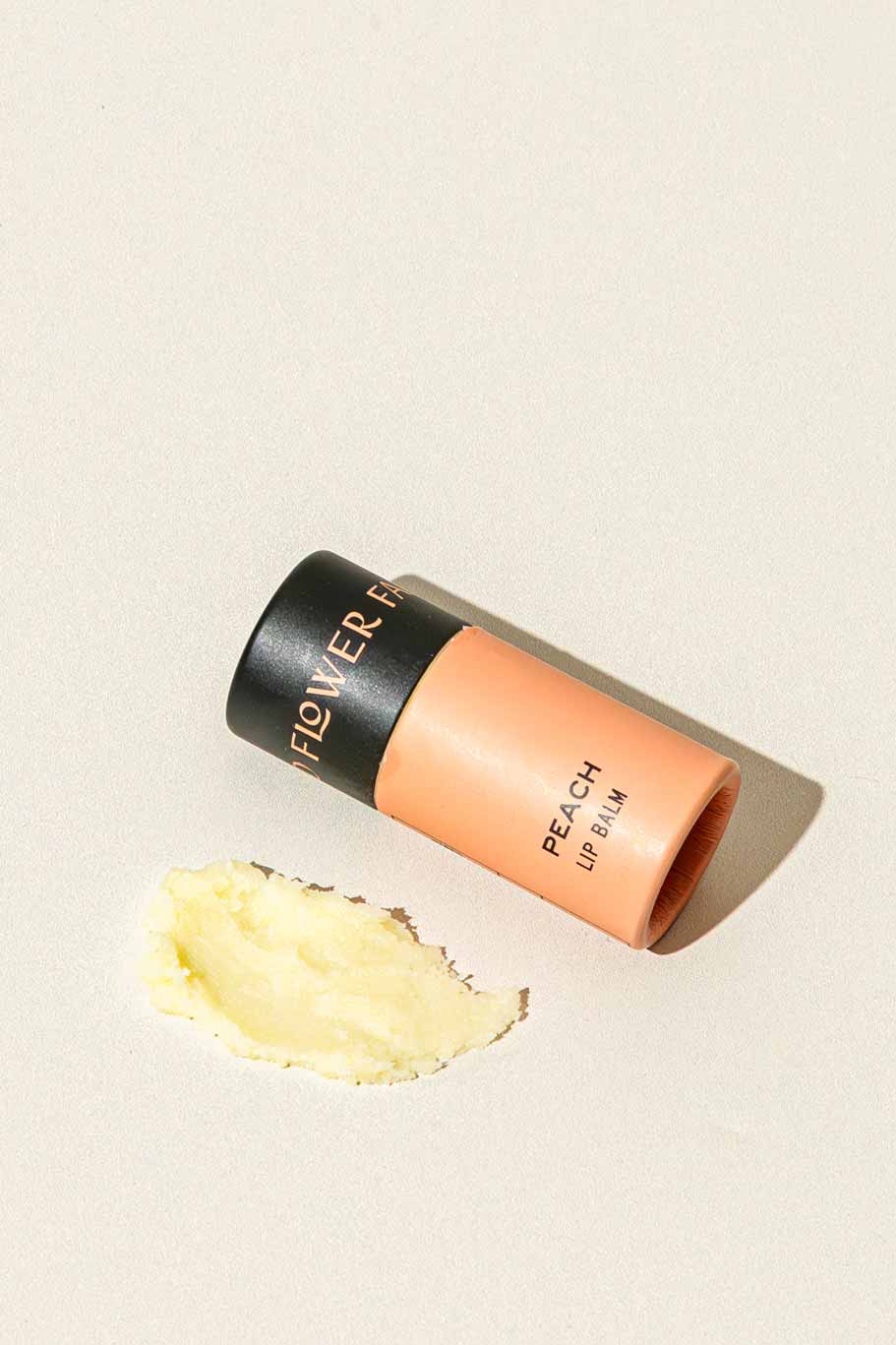 A peach-colored tube of Good Flower Farm Peach Organic Lip Balm lies on its side beside a smear of balm on a light, textured surface.