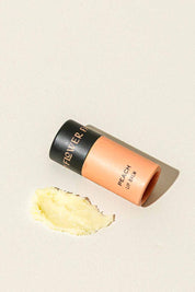 A peach-colored tube of Good Flower Farm Peach Organic Lip Balm lies on its side beside a smear of balm on a light, textured surface.